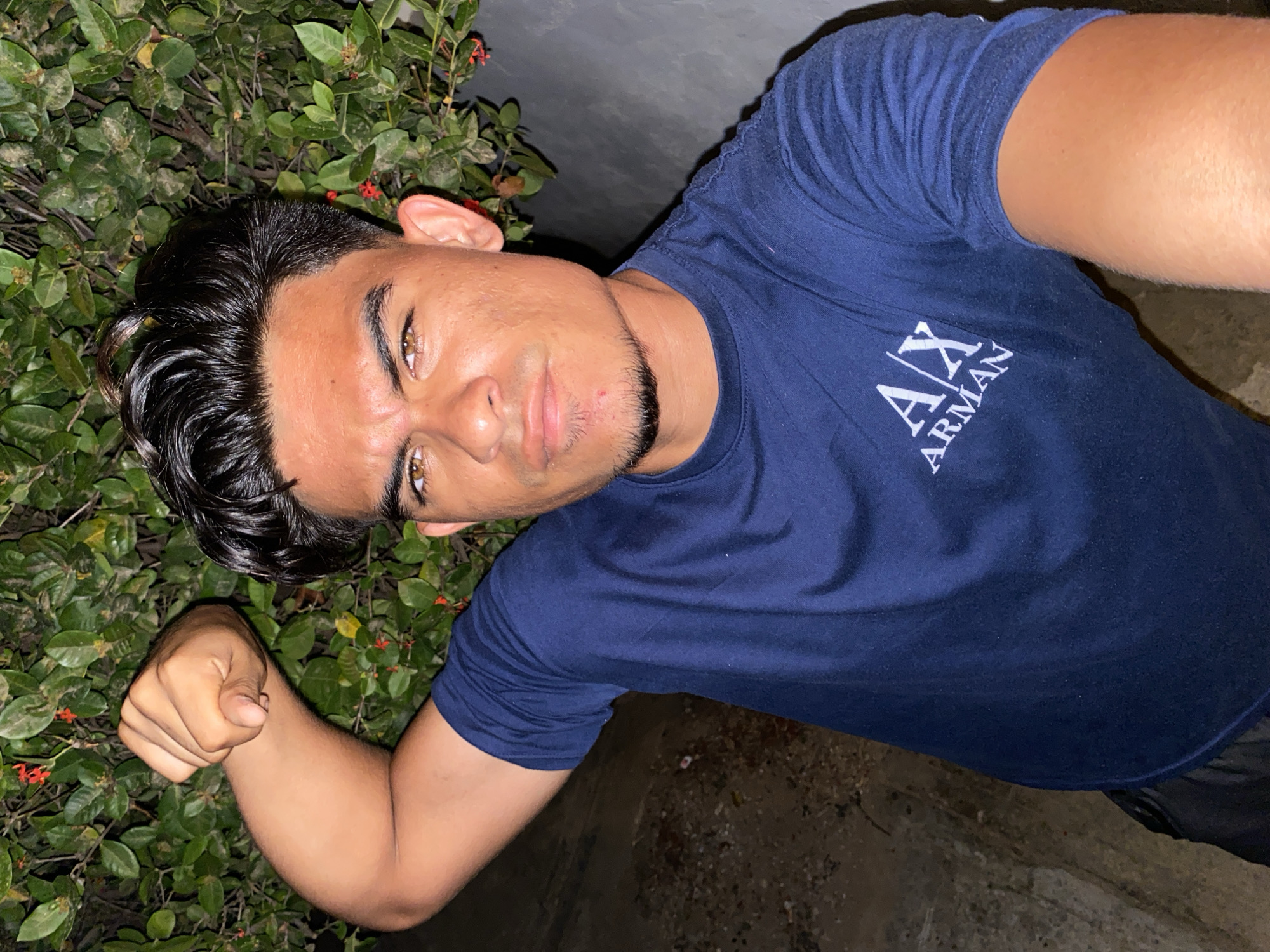 A young man poses in a selfie outdoors, next to dense green foliage with small red flowers. He wears a dark blue t-shirt with a white logo, "A/X ARMANI." His hair is dark, styled back, and he has brown eyes and a light beard. His skin tone is light brown. He flexes one arm, making a fist. The background is a dark, rough surface, possibly concrete. The location is unspecified beyond the generalized area of Juan Griego, Venezuela. The image composition suggests he is standing beside a wall or structure covered in greenery, likely at night or in dim lighting.