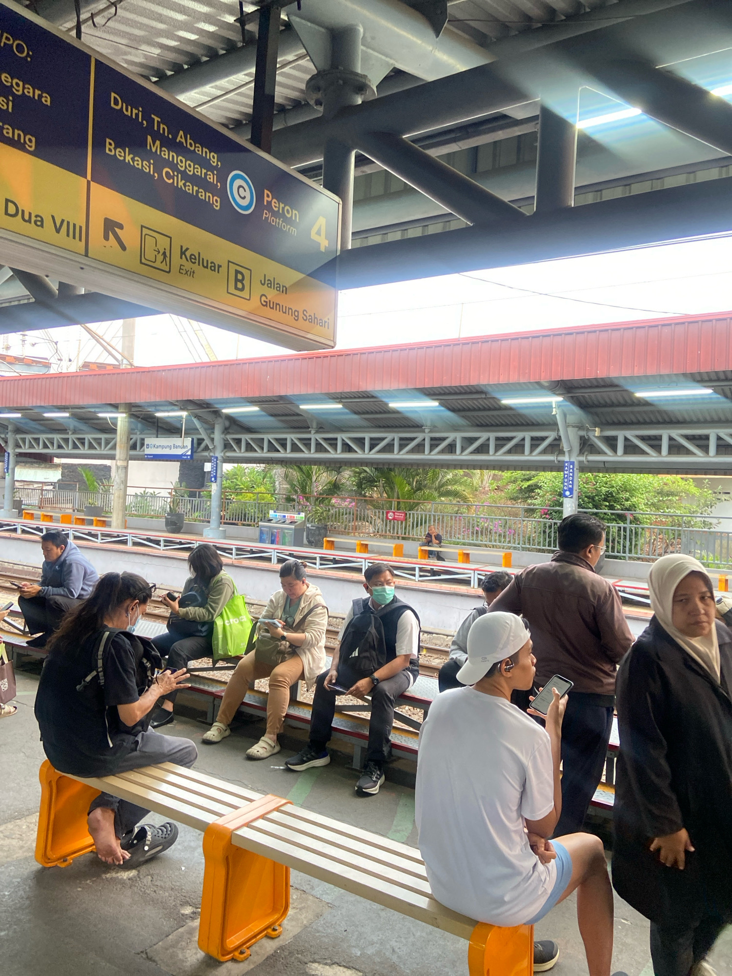 The image depicts a scene at a train station in North Jakarta, Indonesia, likely Cikarang station, as indicated by directional signs. The station features a covered platform with a red roof supported by a metal truss system. Several people are seen waiting on orange-seated benches, some looking at their phones, others sitting with their belongings. The people are a mix of genders and ages, dressed in casual attire. One sign, in Indonesian, directs to "Peron 4" (Platform 4) and lists destinations such as "Duri, Tn. Abang, Manggarai, Bekasi, Cikarang" and "Jalan Gunung Sahari." Another sign indicates "Keluar" (Exit) and "Dua VIII." The ground is paved concrete, and the tracks are visible. The lighting suggests it is daytime, with sunlight streaming through gaps in the roof, creating bright highlights and shadows. The environment appears functional and utilitarian, typical of a public transportation hub.