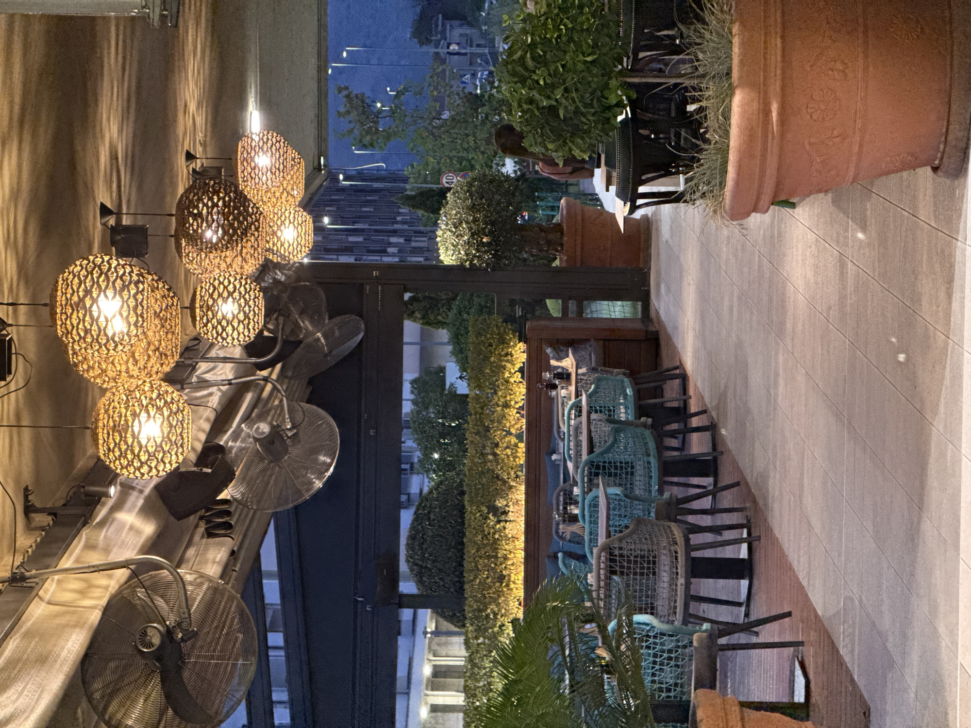 An outdoor dining area at dusk features multiple round, woven pendant lights hanging from the ceiling. Two industrial-style oscillating fans are mounted overhead. The area is furnished with tables and chairs, with some chairs being a light teal color. Large terracotta pots with plants are strategically placed around the seating area. In the background, there are manicured bushes and trees, with buildings and streetlights visible in the distance. The ground is paved with large, light-colored tiles. One person is partially visible in the background.