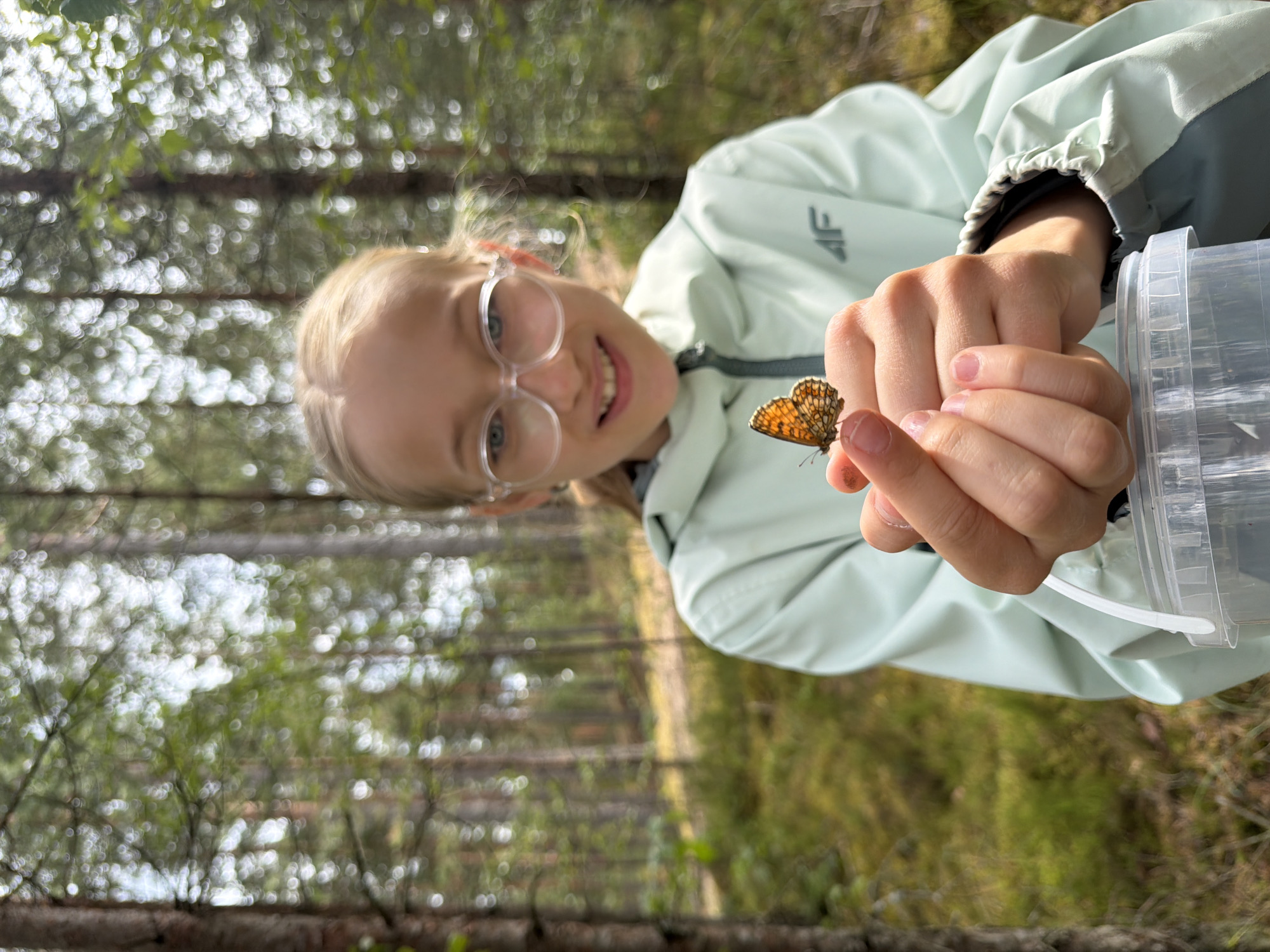 A young girl with blonde hair and clear glasses is holding a small butterfly on her finger in a forest. She is wearing a light green jacket with a grey stripe. The background is a blur of green trees and sunlight. The girl is smiling and looking at the butterfly. A clear plastic container is visible at the bottom of the frame. The scene appears to be set in a natural environment, possibly a park or wooded area, consistent with the description of the city of Mucenieki, Latvia, which is known for its forests and natural landscapes.