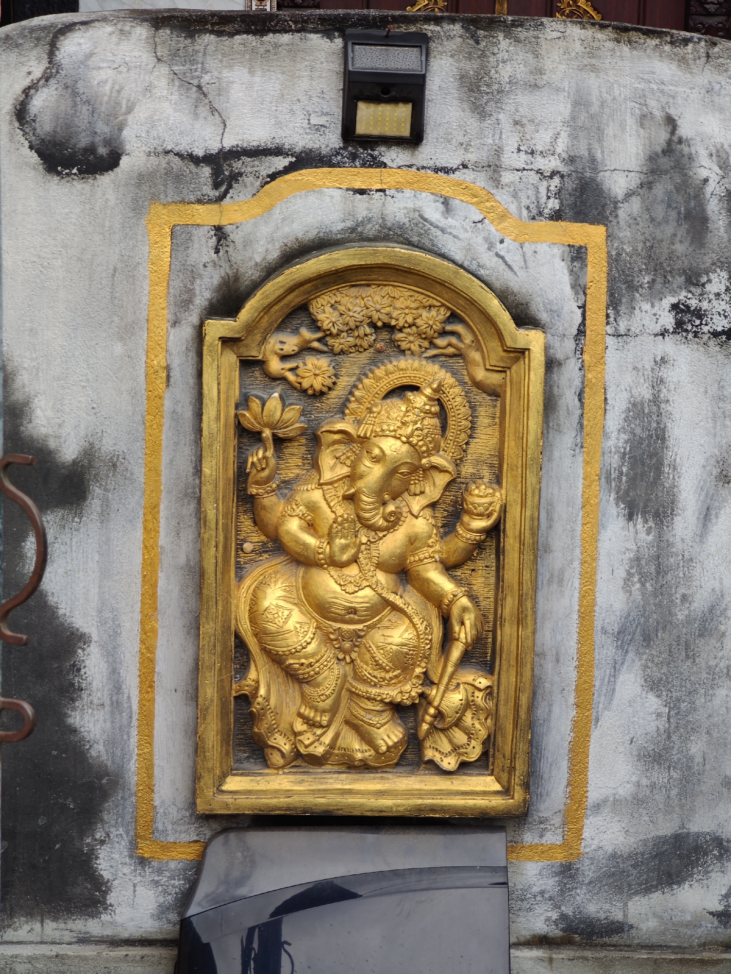 A medium-angle shot depicts a golden relief sculpture of a Hindu deity, possibly Ganesha, mounted on a weathered, gray-and-black concrete wall. The sculpture, encased in a golden-bordered frame, features the deity with multiple arms holding various symbolic objects. A leafy tree is carved above the deity. A black rectangular object is installed above the sculpture on the wall. The wall shows signs of age and weather, with dark stains and cracks. Parts of a dark-colored vehicle are visible at the bottom of the image. The sculpture is located on Nusa Penida, Indonesia.