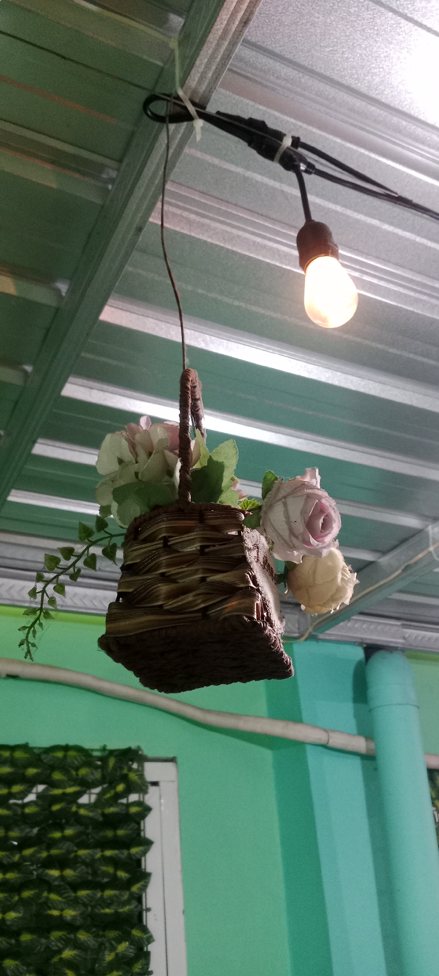The image shows a hanging basket filled with artificial flowers suspended from a metallic ceiling. A light bulb is illuminated nearby, also hanging from the ceiling. The background features a light green wall. The scene appears to be an indoor or covered outdoor space, possibly a shop or a garden area. There is no indication of the specific location within the city of Tuban, Indonesia. The image does not contain any people.