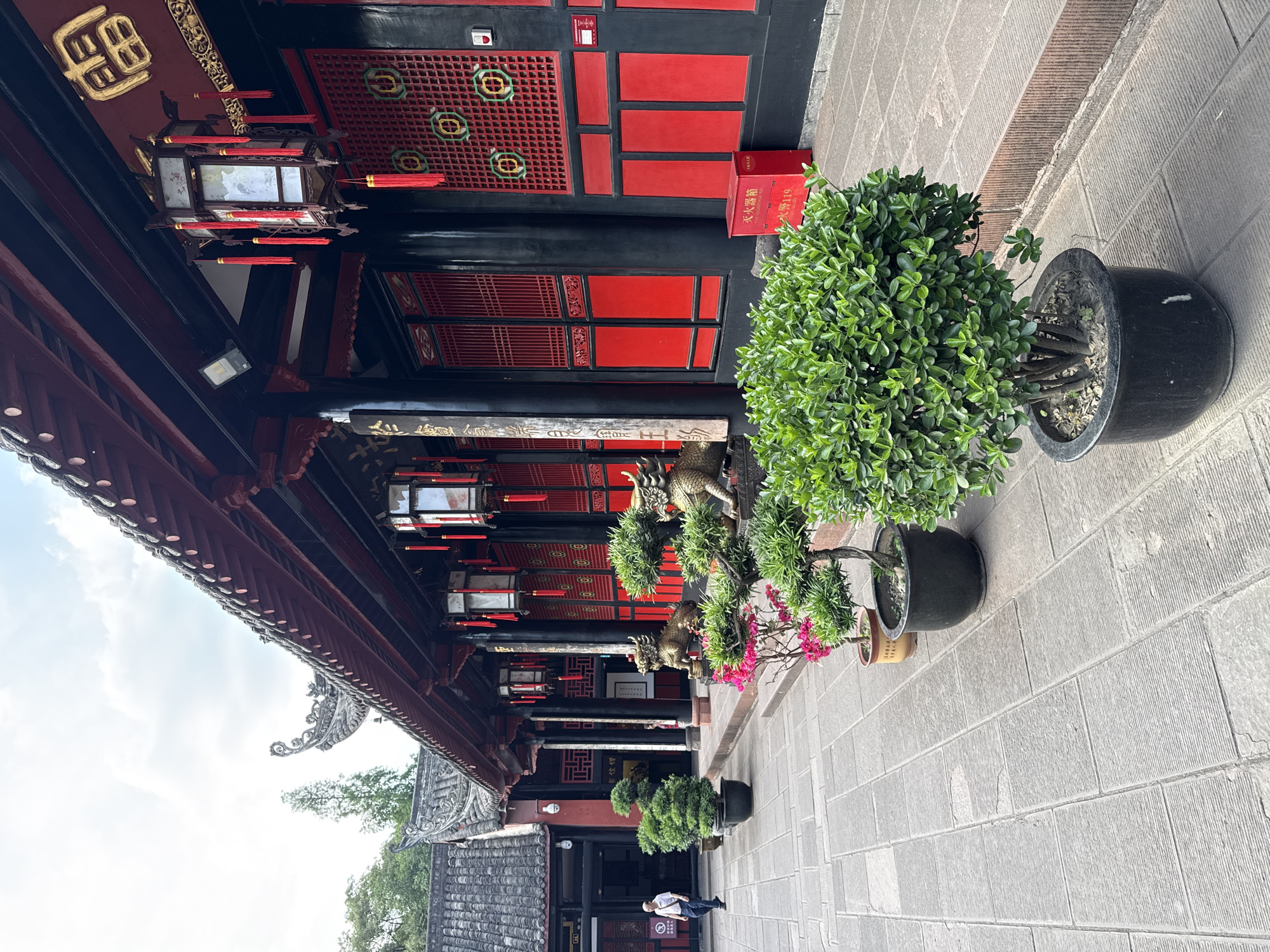 The photograph depicts an exterior view of a traditional Chinese building located in the Qingyang District of China. The building has a red and black color scheme, with intricate lattice work, traditional lanterns, and decorative eaves. Several potted plants line the walkway adjacent to the building. A person is standing near the entrance of the building in the background. The walkway is paved with stone tiles. The sky is visible in the upper left corner of the image.