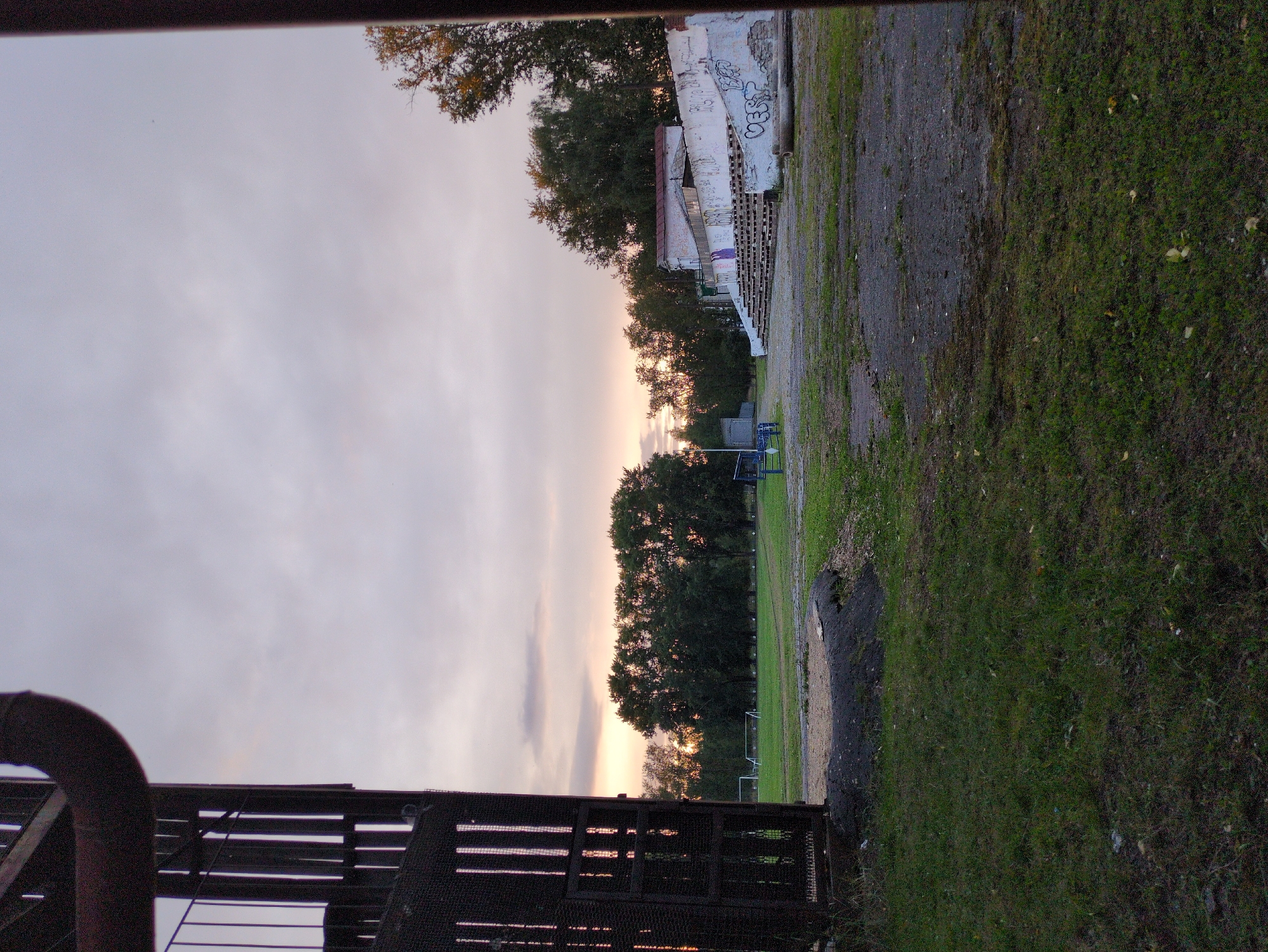 The image shows an outdoor view of a sports field at dusk. In the foreground, on the left, is a dark, industrial-looking structure with vertical metal bars and a large, curved pipe extending from the top left. The ground in the foreground is covered with patchy grass and dark, rough terrain.
In the midground, a grassy field is visible with several white soccer goals. Beyond the field, a line of dense green trees separates the field from the sky. To the right, there is a concrete structure with tiered seating, appearing to be an old stadium grandstand. The wall of the grandstand is covered in graffiti. A small building is situated behind and above the grandstand.
The sky is filled with a pale, cloudy twilight, with hints of orange and pink light near the horizon, suggesting the sun has recently set. The overall scene appears to be an abandoned or disused sports facility in a rural or semi-urban setting. The location is identified as Kulebaki, Russia. There are no people visible in the image.