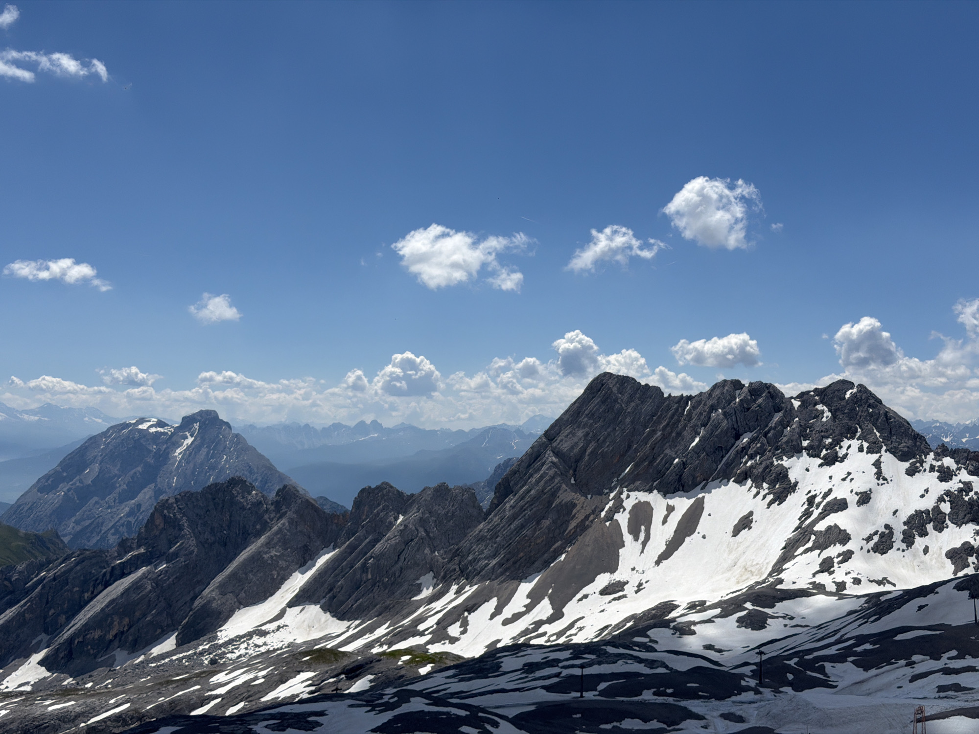 The image displays a panoramic view of a mountainous landscape under a bright blue sky with scattered white clouds. In the foreground, rugged, dark gray rock formations are partially covered by patches of white snow, suggesting a high-altitude environment. The slopes are steep and uneven, with some areas appearing rocky and barren while others are blanketed in snow.
In the mid-ground, more mountain peaks are visible, with their jagged ridges silhouetted against the sky. The distant background features a range of mountains fading into a hazy blue, indicating significant depth and distance. One prominent peak in the mid-left of the image is particularly large and imposing, with snow capping its summit and parts of its slopes.
The lighting suggests it is daytime, with clear visibility. The scene is devoid of any human presence, animals, or man-made structures, except for what appears to be a few ski lift towers or poles scattered sparsely across the snowy slopes in the lower right portion of the image. The overall environment is a natural, alpine landscape.
This mountainous scene is consistent with the landscape found in and around Garmisch-Partenkirchen, Germany, a region known for its prominent Bavarian Alps, including the Zugspitze, Germany's highest peak, which features similar dramatic rocky peaks and snowfields.