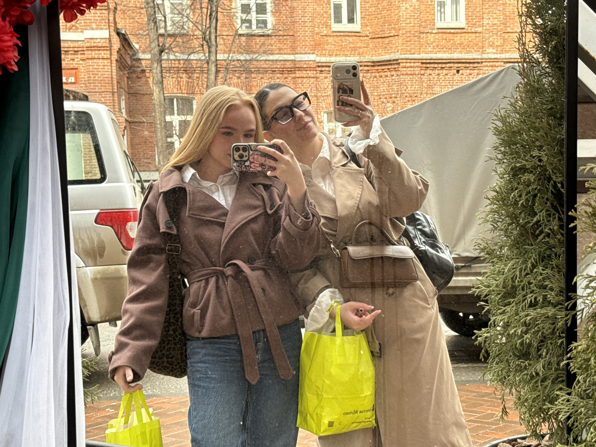 Two young women are taking a selfie in front of a mirrored surface. The woman on the left has long, straight blonde hair and is wearing a brown coat tied at the waist with a matching belt. She is also wearing a denim skirt or jeans and a leopard-print shoulder bag. She holds a yellow shopping bag in her left hand and a smartphone in her right, using it to take the picture. The woman on the right is wearing a beige trench coat, black-framed glasses, and her hair is pulled back. She holds a smartphone in her right hand, also using it to take the picture. Both women are wearing white collared shirts under their coats. They are standing in front of a reflective surface, likely a shop window, which shows a reflection of a brick building, trees, a silver SUV, and a covered vehicle. A green bush is visible on the right side of the frame. There are white curtains on the left side of the frame. The city is Perm, Russia.