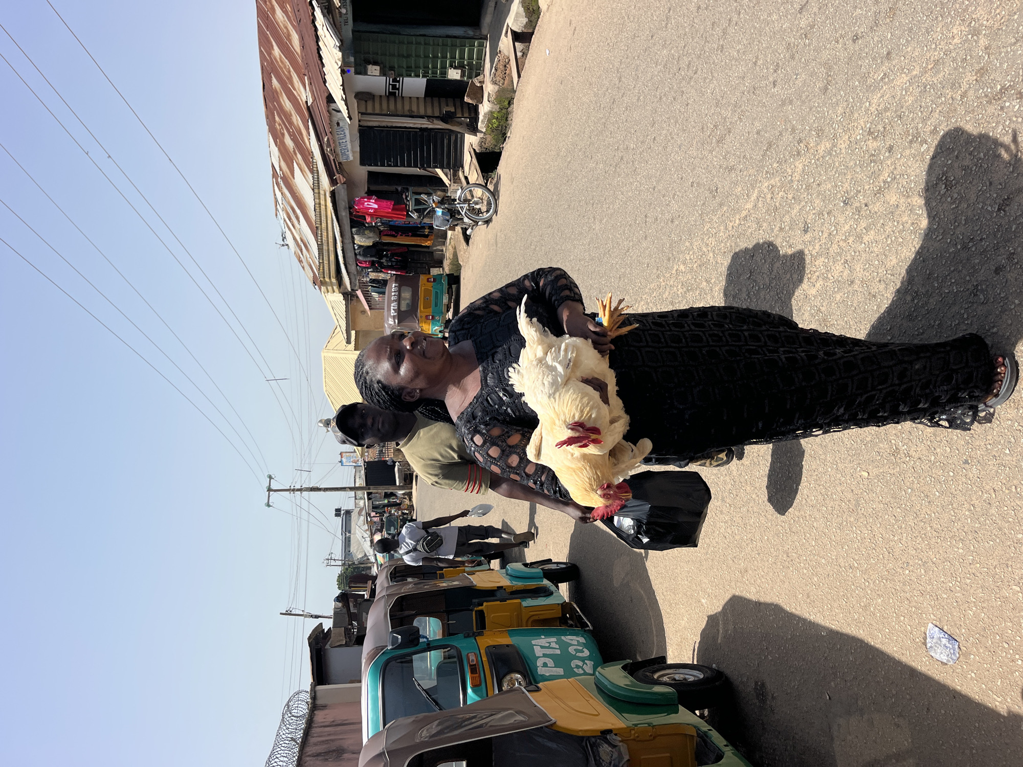 In an outdoor scene in an unknown city, a woman in a black dress with an intricate pattern carries a live, light-colored chicken in her arms. She also holds a black plastic bag in her left hand. To her left, a row of colorful auto-rickshaws, with the text "PTA 204" visible on one, are parked on the side of an asphalt road. Behind the woman, a man in a green shirt and a cap walks away from the camera, carrying a white object and a dark bag. In the background, shops with corrugated metal roofs line the street, displaying clothing and other goods. A motorcycle is parked in front of one of the shops. The sky is clear and blue, and the sun casts strong shadows on the ground. The environment appears to be a bustling street market or commercial area.