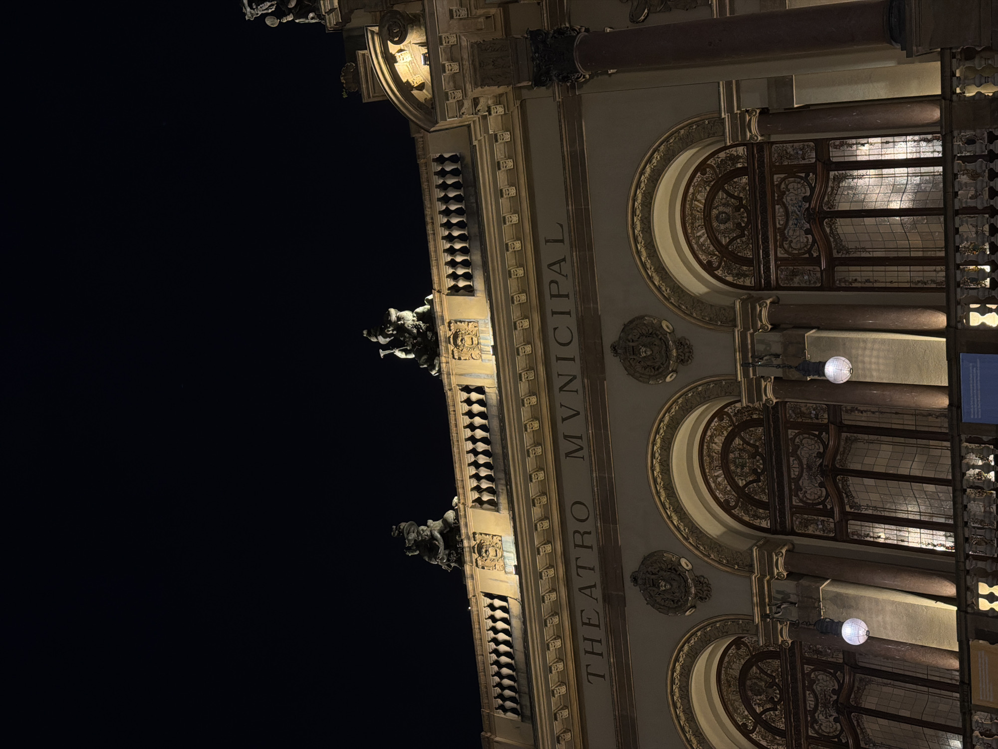The image shows the facade of the Theatro Municipal de São Paulo, Brazil at night. The building is illuminated, highlighting its ornate architecture. The top of the building features sculptures and decorative railings against a dark night sky. Below this, the name "THEATRO MUNICIPAL" is inscribed in large letters. Further down, arched windows with intricate stained glass designs are visible, flanked by columns. Hanging lights illuminate the facade. The overall impression is one of grandeur and historical significance.