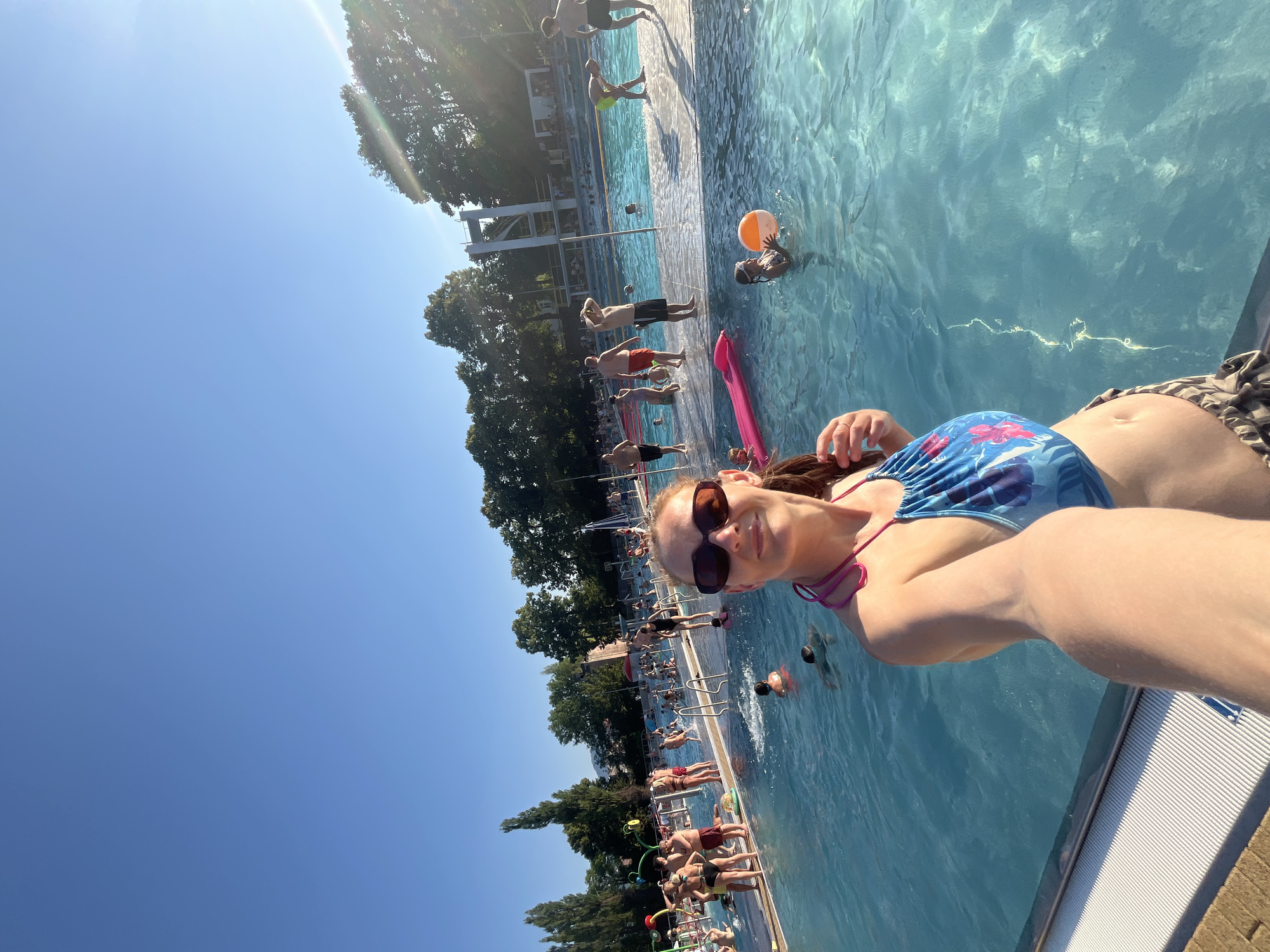An outdoor public swimming pool on a sunny day. A woman in a blue and pink swimsuit is taking a selfie in the foreground. She is wearing sunglasses and has her hair pulled back. The pool is filled with blue water and many people are swimming and sunbathing. There are trees and buildings in the background.