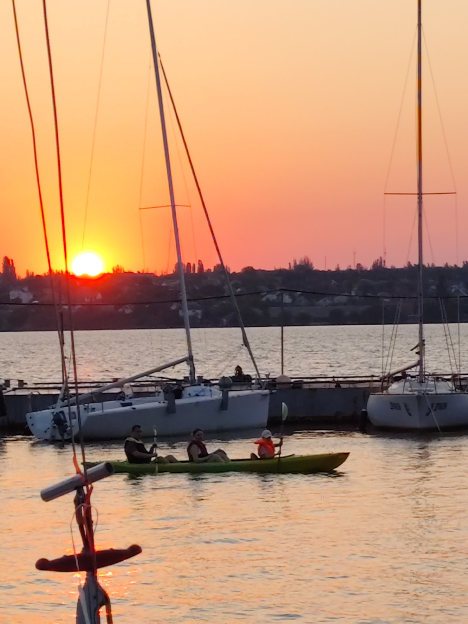 The media file captures a serene sunset over a body of water, likely the Southern Bug river in Mykolaiv, Ukraine. Silhouetted against the vibrant orange and pink sky are several sailboats docked at a pier. In the foreground, a green kayak is being paddled by three individuals. The two individuals in the rear are adults, one wearing a yellow life vest and the other a red one. The person in the front appears to be a child wearing an orange life vest. The water reflects the warm hues of the sunset, creating a peaceful and tranquil atmosphere. The time of day is clearly sunset, indicated by the low position of the sun and the intense colors in the sky. The weather appears calm and pleasant, suitable for water activities. The presence of sailboats and a dock suggests a marina or harbor setting. The overall mood is one of relaxation and enjoyment of nature's beauty. There is no visible text in the image.