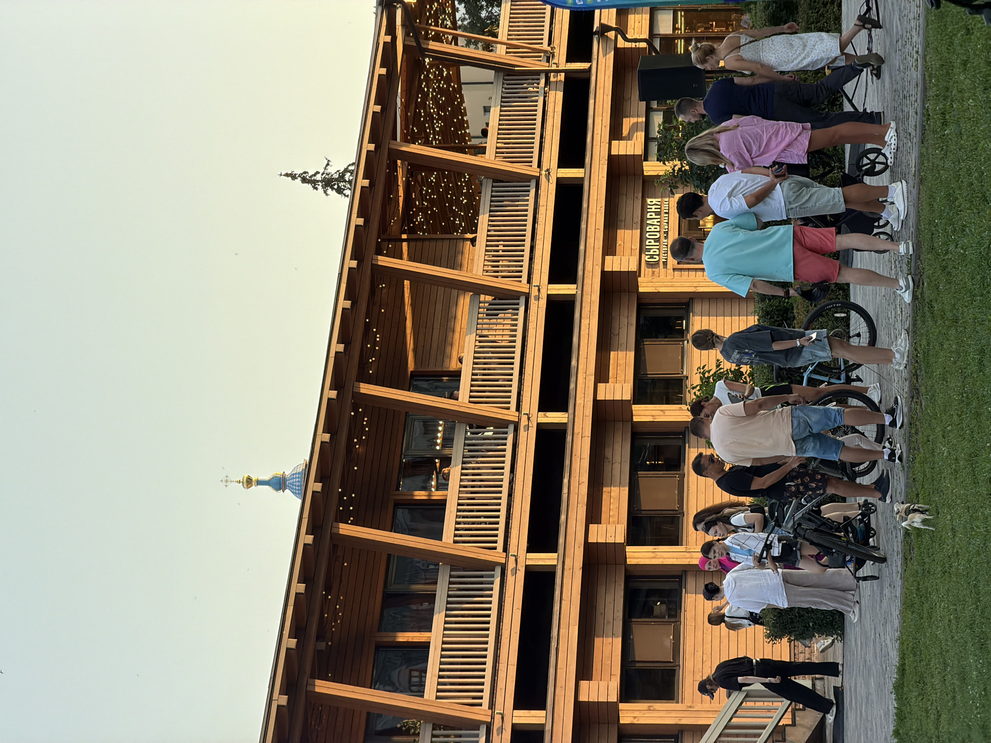 The image captures a group of people gathered outside a wooden building in Kazan, Russia, possibly in the late afternoon or early evening given the lighting. The building has multiple levels with balconies, adorned with string lights. A sign above a ground-floor entrance reads "СЫРОВАРНЯ" (Syrovarnya), which translates to "Cheese Dairy" or "Fromagerie." In the background, a blue-domed church steeple with a golden cross is visible against a pale sky. A few birds are seen flying in the distance.
In the foreground, several individuals are standing around, some interacting, while others are looking away or engaged with their phones. Two bicycles are parked near the group. A small dog is visible on the grass in the lower-left portion of the frame. The ground is paved with interlocking stones, and a grassy area is present on the left. The overall atmosphere appears casual and social.
