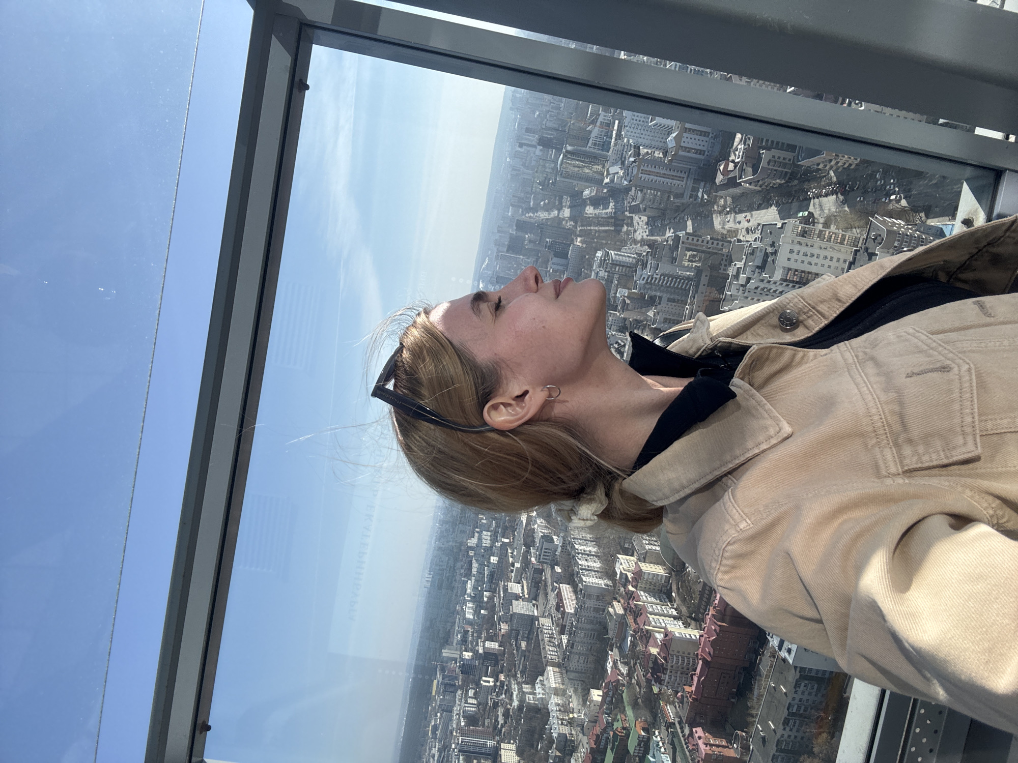A young woman stands indoors in a high-rise building, possibly a skyscraper. She is looking up at a light source, possibly the sun. She wears a beige jacket over a dark top, and sunglasses are tucked into her hair. The background, visible through a large window, shows a dense urban landscape of Yekaterinburg, Russia, with numerous buildings.
