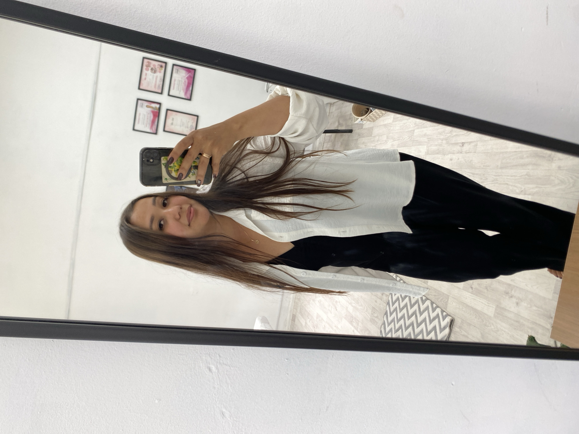 A young woman is taking a mirror selfie in a room. She is wearing a black slip dress and an open white button-down shirt over it. She has long, brown hair and is wearing a necklace. She is holding a smartphone in her right hand and has a ring on her left ring finger. The room has white walls and a light wood floor. In the background, there are several framed certificates on the wall. A rug with a zig-zag pattern is visible on the floor. The scene appears to be located in a home or a business establishment. The image was taken in Akyar, Russia.