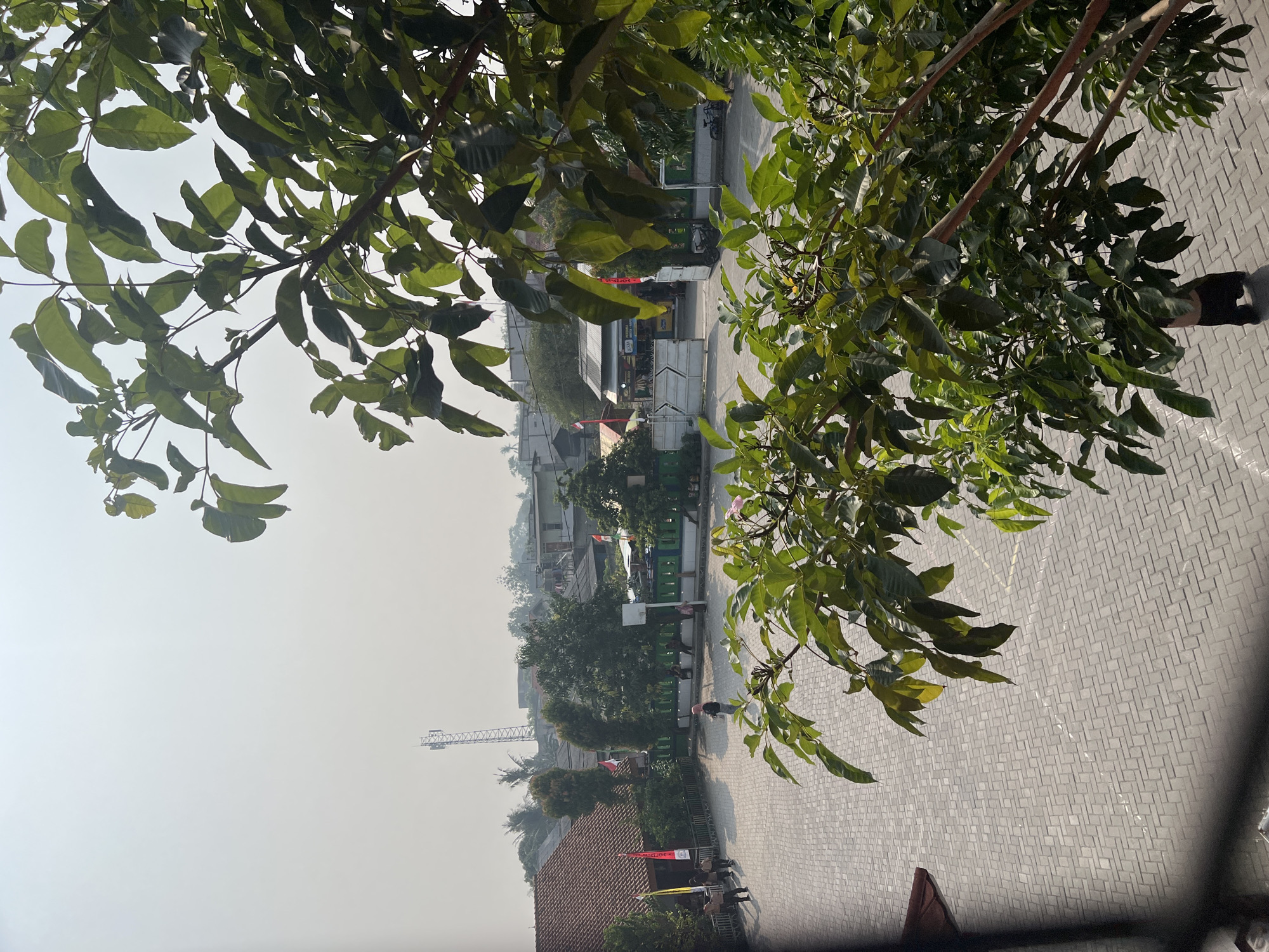This is an outdoor, eye-level medium shot of a schoolyard in Gelam Jaya, Indonesia, on a bright, hazy day. In the foreground, the branches of a lush green tree with broad leaves obscure the upper right portion of the image. The ground is paved with light gray interlocking bricks arranged in a herringbone pattern.

The schoolyard appears to be empty except for a few people. On the left side, a group of three children in what appears to be school uniforms are walking. In the center of the frame, a person wearing a pink headscarf and a dark shirt is walking across the yard. Further back, near a green fence, several people are seated.

In the background, the school buildings have red-tiled roofs and cream-colored walls. Several Indonesian flags are displayed, including some on flagpoles. Beyond the school, a dense urban landscape with various buildings and trees is visible, and a tall communication tower stands prominently in the distance. The overall impression is a typical school scene in a developing urban environment.
