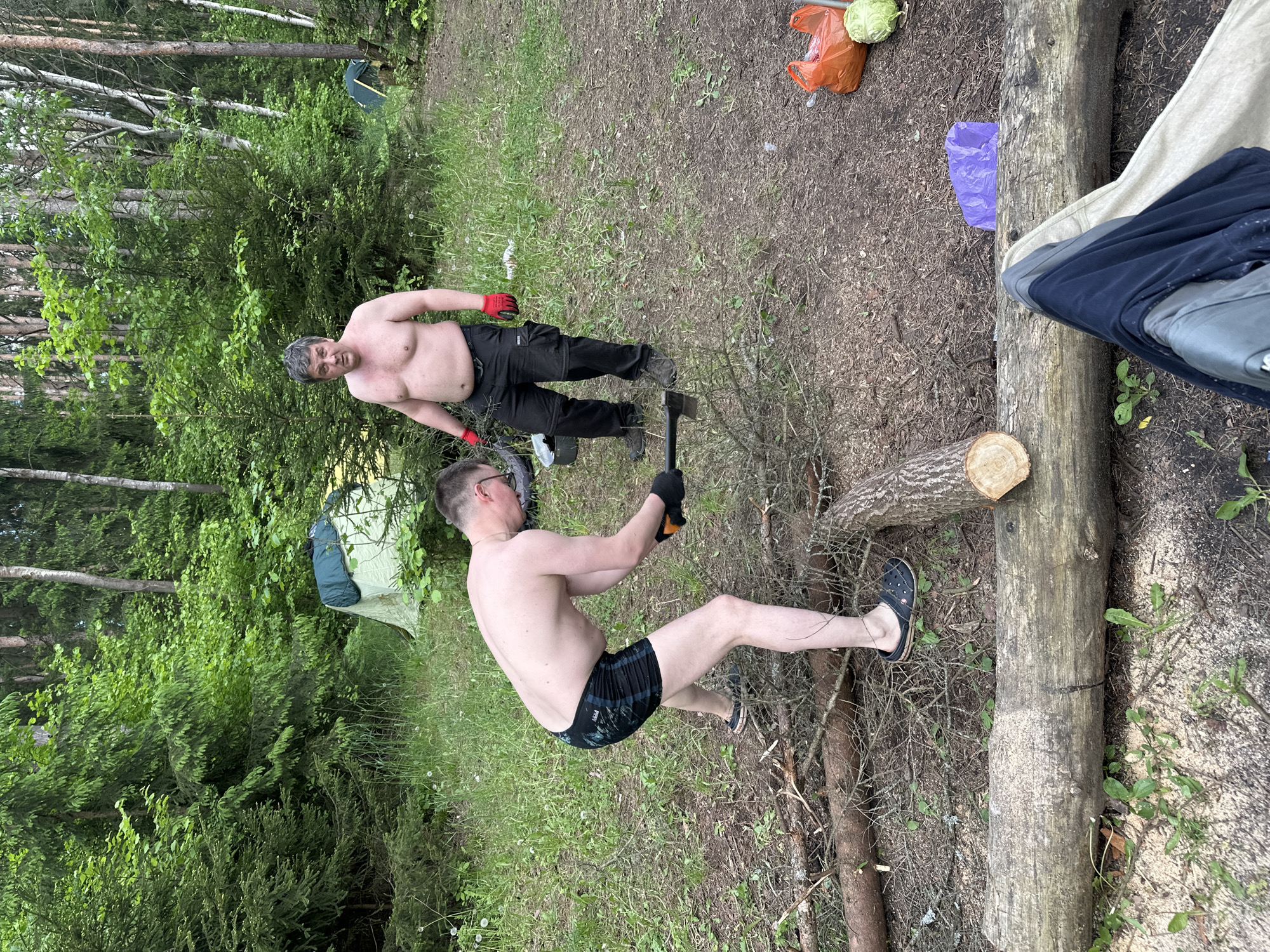 The scene is outdoors in a wooded area of Папярнянскі сельскі Савет, Belarus. Two shirtless men are engaged in firewood preparation. One man, wearing black pants and red gloves, stands in the background. The other man, clad in shorts, glasses, and black gloves, wields an axe to split a log. A green tent is set up in the background amidst the trees. A cut log lies on the ground to the right, alongside a larger log that appears to be in use as a work surface. A cabbage and some trash are also visible in the background.
