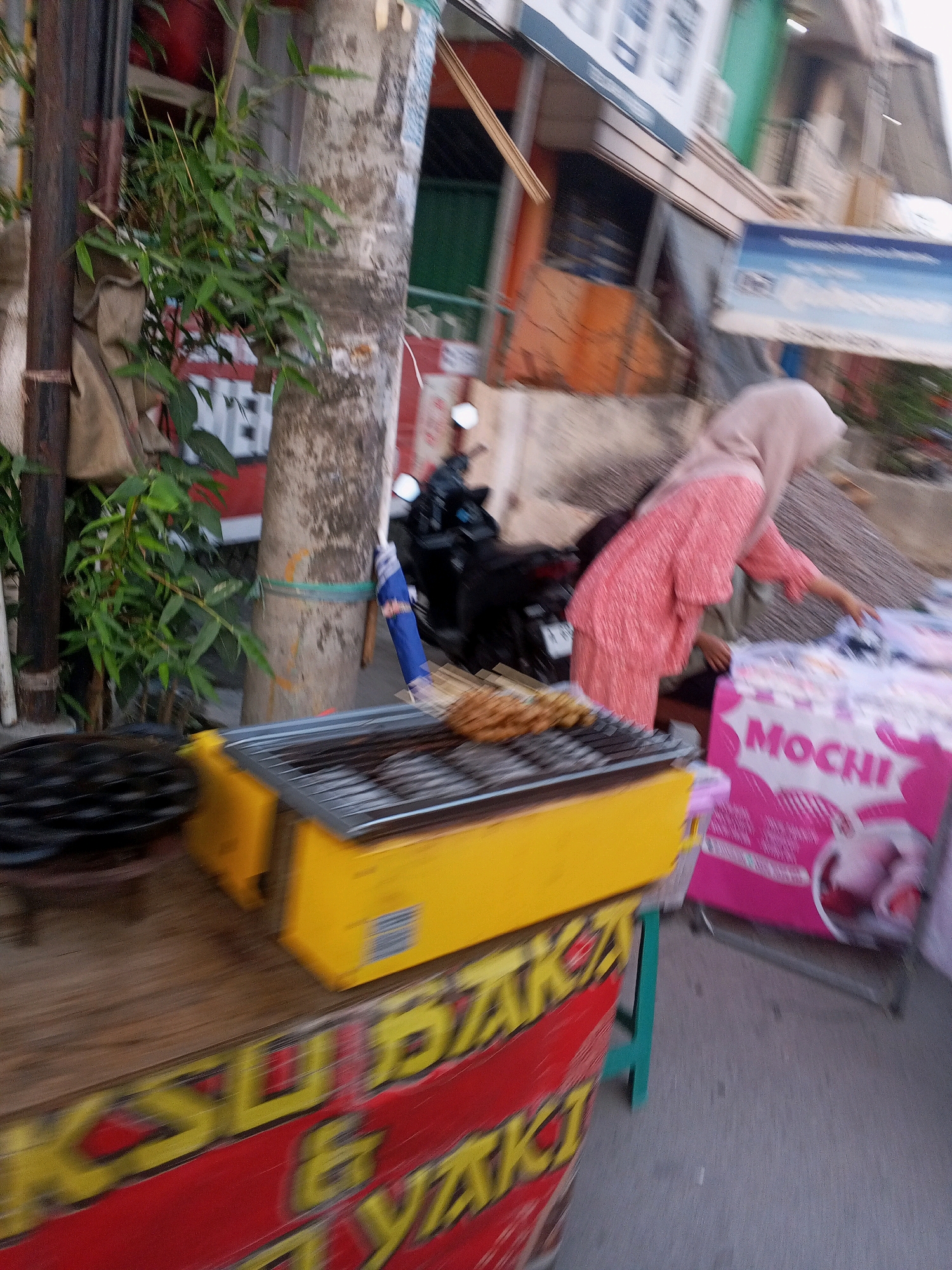 In Ciruas, Indonesia, a street scene unfolds with a focus on a food stall. A woman, partially obscured and wearing a pink hijab and a textured pink garment, appears to be serving or arranging items at the stall. In the foreground, a yellow grill sits on a wooden surface, with what appears to be food on skewers resting on the grill. To the left, a black grill pan is visible. A large red and yellow sign with text is also present, although the writing is blurred. Behind the stall, a motorcycle is parked. The environment suggests an outdoor setting, likely a street or market.