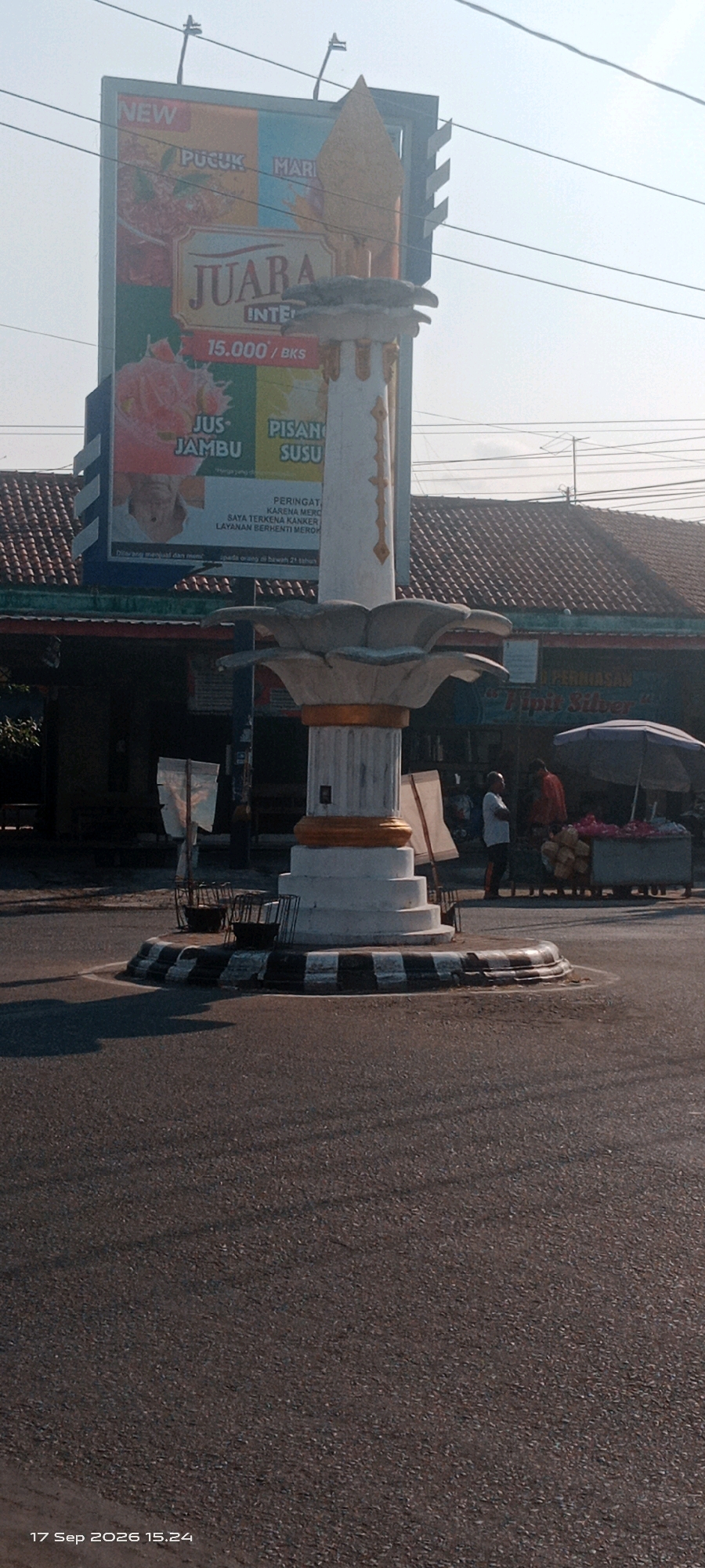 The image shows a traffic circle in Wedi, Indonesia, featuring a central monument with a large billboard behind it. The monument is white with gold accents and has a lotus-like structure at the top. The billboard advertises "JUARA" products, including "PUCUK", "MARI", "JUS JAMBU", and "PISANG SUSU", with a price of "15.000/BKS". There is also a health warning on the billboard about smoking.
In the background, a two-story building with a tiled roof is visible, likely shops or stalls. Several people are present: two men are standing near a cart selling what appears to be flowers or produce, and other figures are indistinct in the background. The scene is set during the daytime, with a clear sky and sunlight casting shadows on the asphalt road. The overall impression is a busy street corner or intersection in a town.