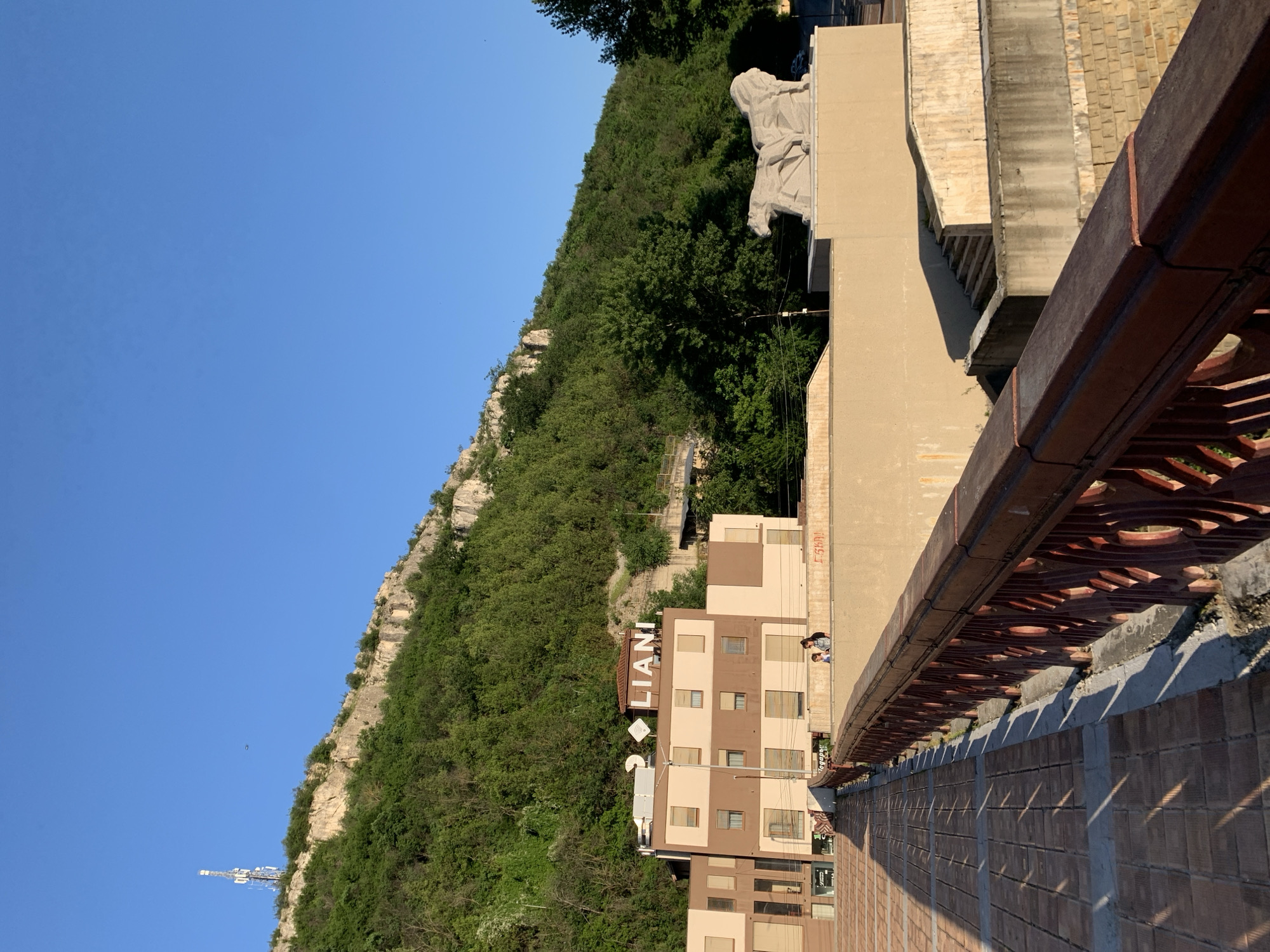 The image presents a view of a city location, specifically showcasing a tall building with the sign "LIANI" prominently displayed. The building is adjacent to a steep, green hillside or mountain. Above the building, on a higher elevation, is a sculpture or monument.
Two individuals are visible near the building's edge, possibly engaging in observation or conversation. The foreground includes a detailed view of a decorative brick structure or wall with intricate patterns. The overall environment includes a clear blue sky and suggests an urban setting integrated with natural features.