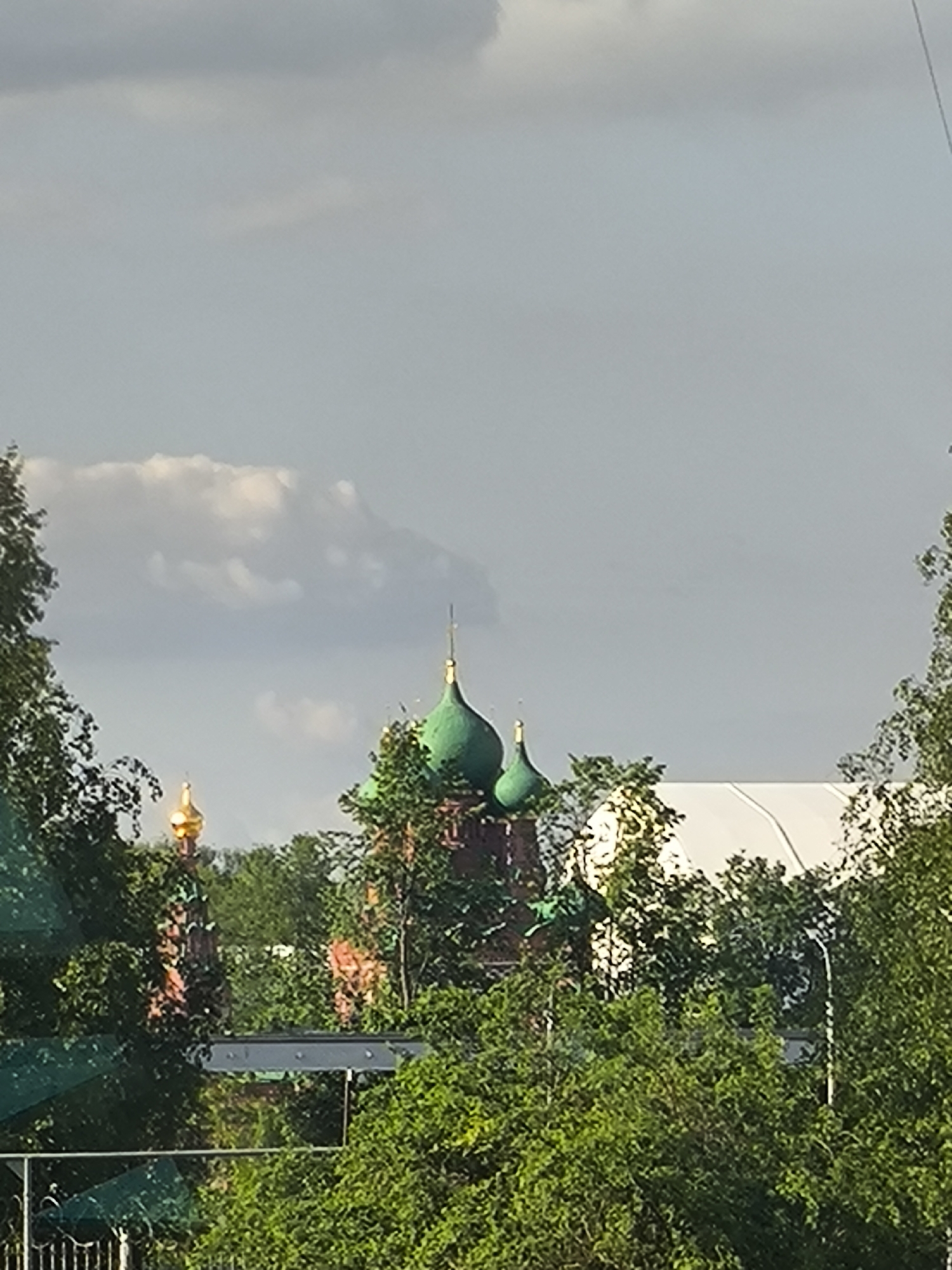 The image shows a view of Moscow, Russia, with several Russian Orthodox churches visible through trees. The church in the center has green domes and a golden cross on top. Another church on the left side has a gold onion dome. A white tent-like structure can be seen behind the trees to the right of the central church. The sky is overcast with gray clouds.