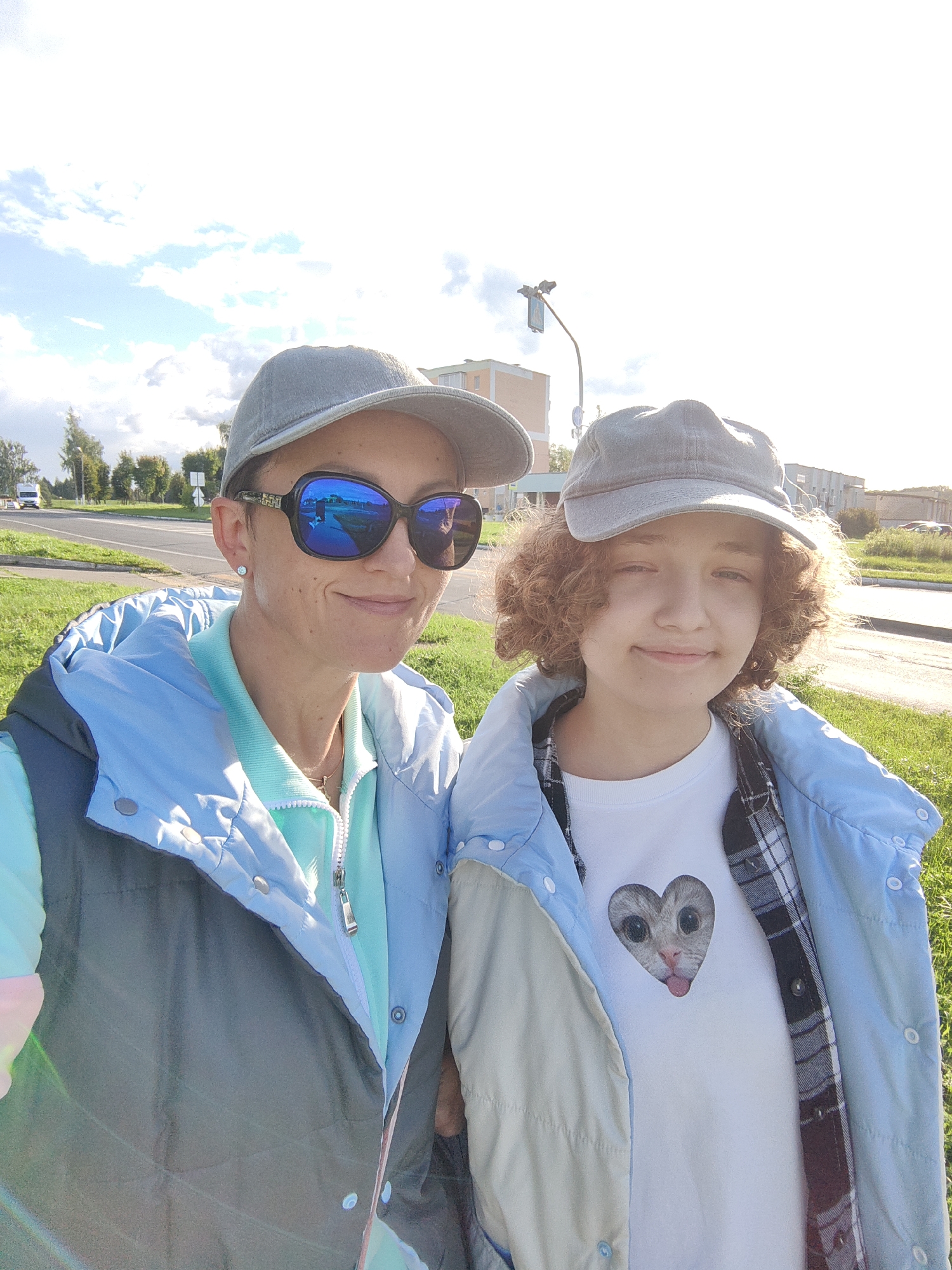 Two individuals, an adult female and a younger person, are taking a selfie outdoors on a paved road with a grassy verge. The adult female on the left is wearing a grey baseball cap, large sunglasses, and a light blue and grey vest over a light blue zippered shirt. The younger person on the right is wearing a grey baseball cap and a white t-shirt with a heart-shaped graphic of a cat with its tongue out, layered under a light blue and tan jacket over a plaid shirt. The background shows a road with a white vehicle visible in the distance, some trees, and buildings. The sky is partly cloudy with bright sunlight. The location is identified as the city of Myadzyel, Belarus.