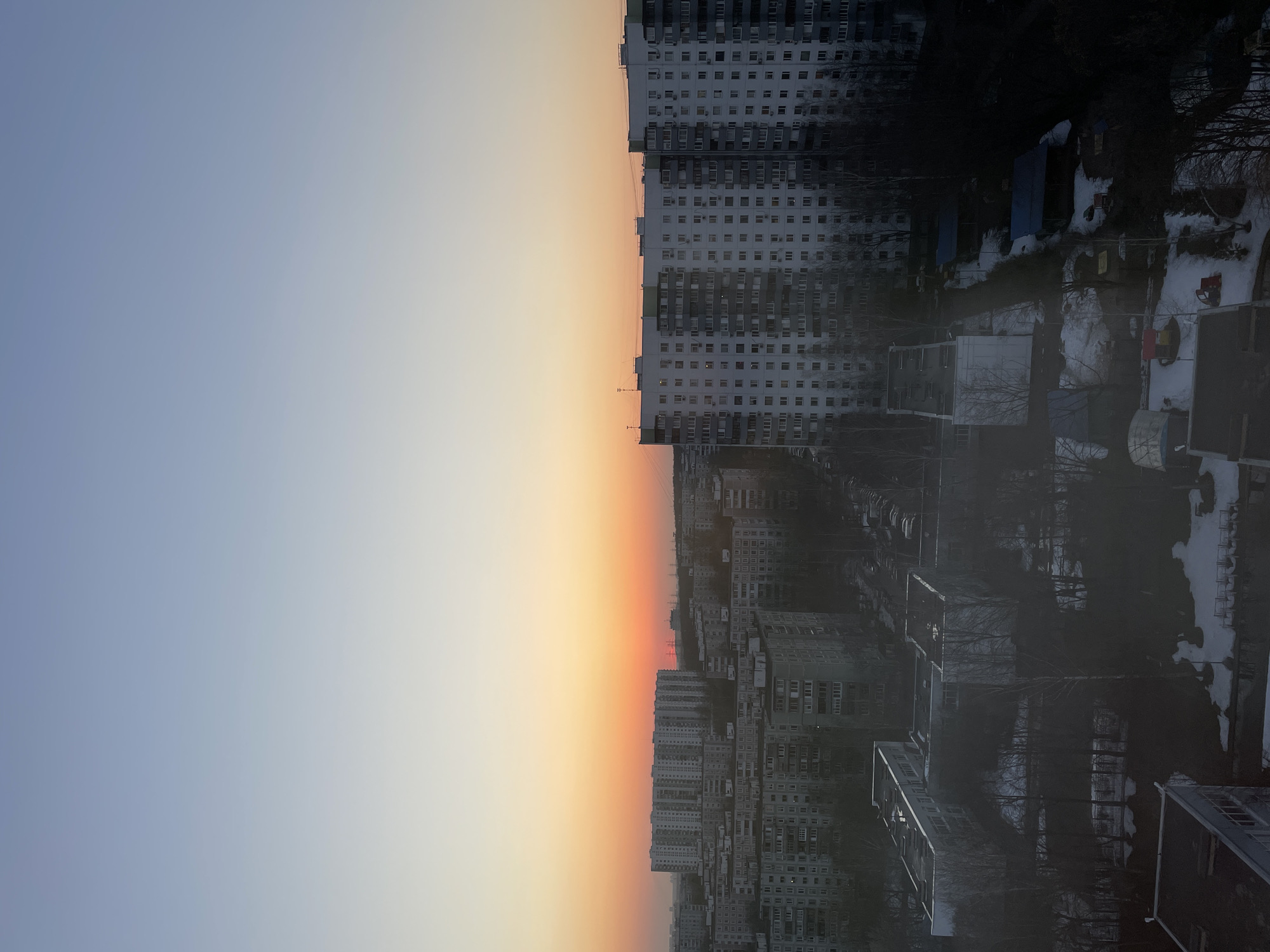 An elevated view captures a sprawling urban residential district during a sunrise or sunset. The sky transitions from a light blue overhead to a vivid orange and red band along the horizon, where the sun's glow is most intense. The landscape is dominated by numerous multi-story apartment buildings, characteristic of Soviet-era "panelka" architecture, with uniform window patterns and light-colored facades. Some buildings feature balconies and external air conditioning units.
In the foreground, partial rooftops of smaller, darker buildings are visible, alongside patches of snow on the ground and numerous bare deciduous trees, indicating a late winter or early spring setting. Interspersed between the larger apartment blocks are areas with parked vehicles. In the far distance, against the bright horizon, silhouettes of high-voltage power lines and towers are discernible. No individual people or specific actions are clearly visible within the frame. This scene depicts a typical peripheral residential area within the city of Moscow, Russia, characterized by its extensive development of mass-produced apartment complexes.