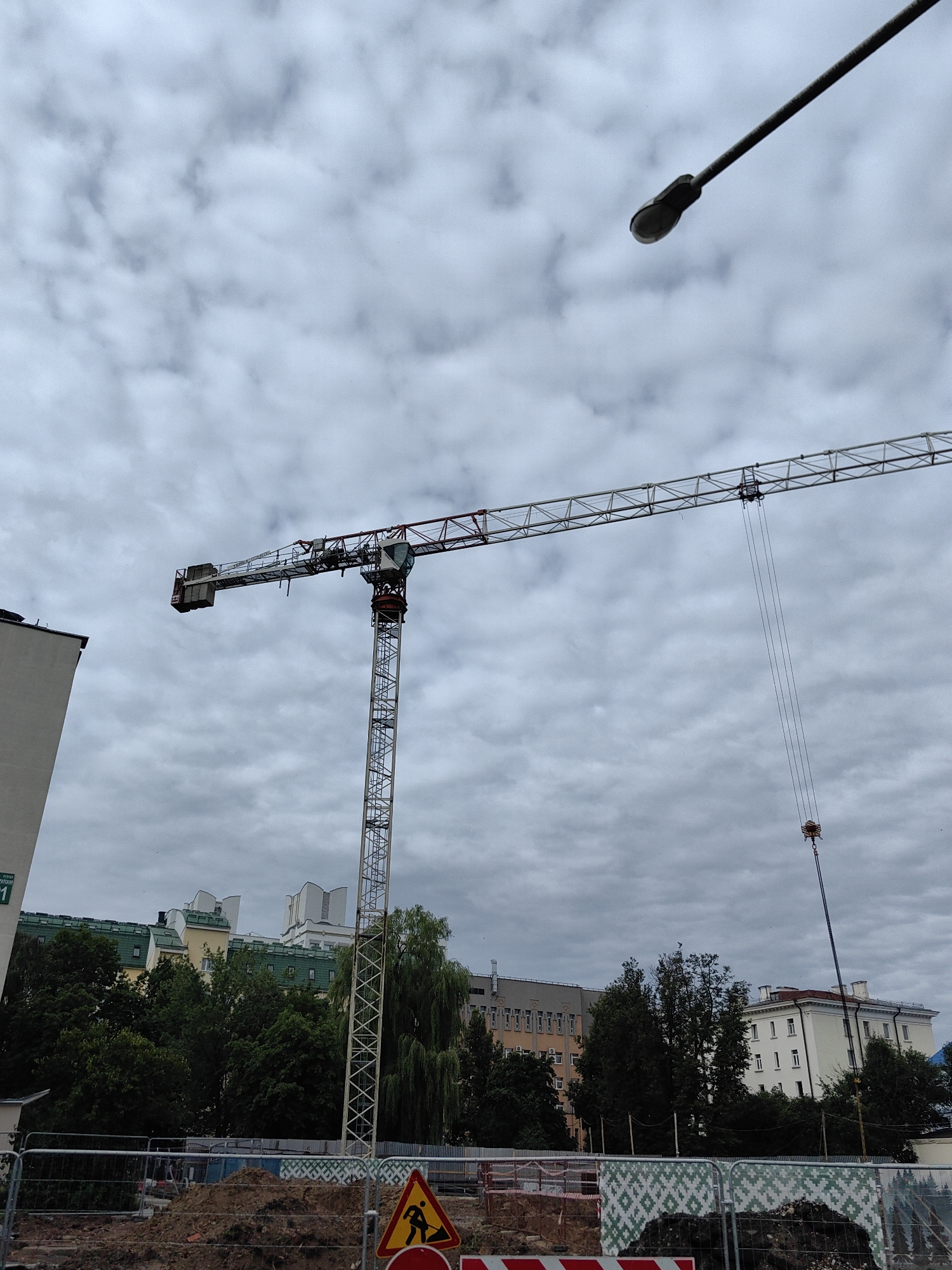 The image depicts a construction site under a cloudy sky in Minsk, Belarus. A tall tower crane dominates the scene, with its long horizontal jib extending across the frame. The crane is a metallic structure, painted white with red accents, and it appears to be in operation, with cables and a hook hanging from the jib. To the left of the crane, a portion of a modern apartment building is visible. In the background, other buildings and trees are present, suggesting an urban setting. The foreground shows construction materials, including piles of dirt, fencing, and a warning sign depicting a person with a shovel. The overall atmosphere is one of ongoing construction and development in a city environment.