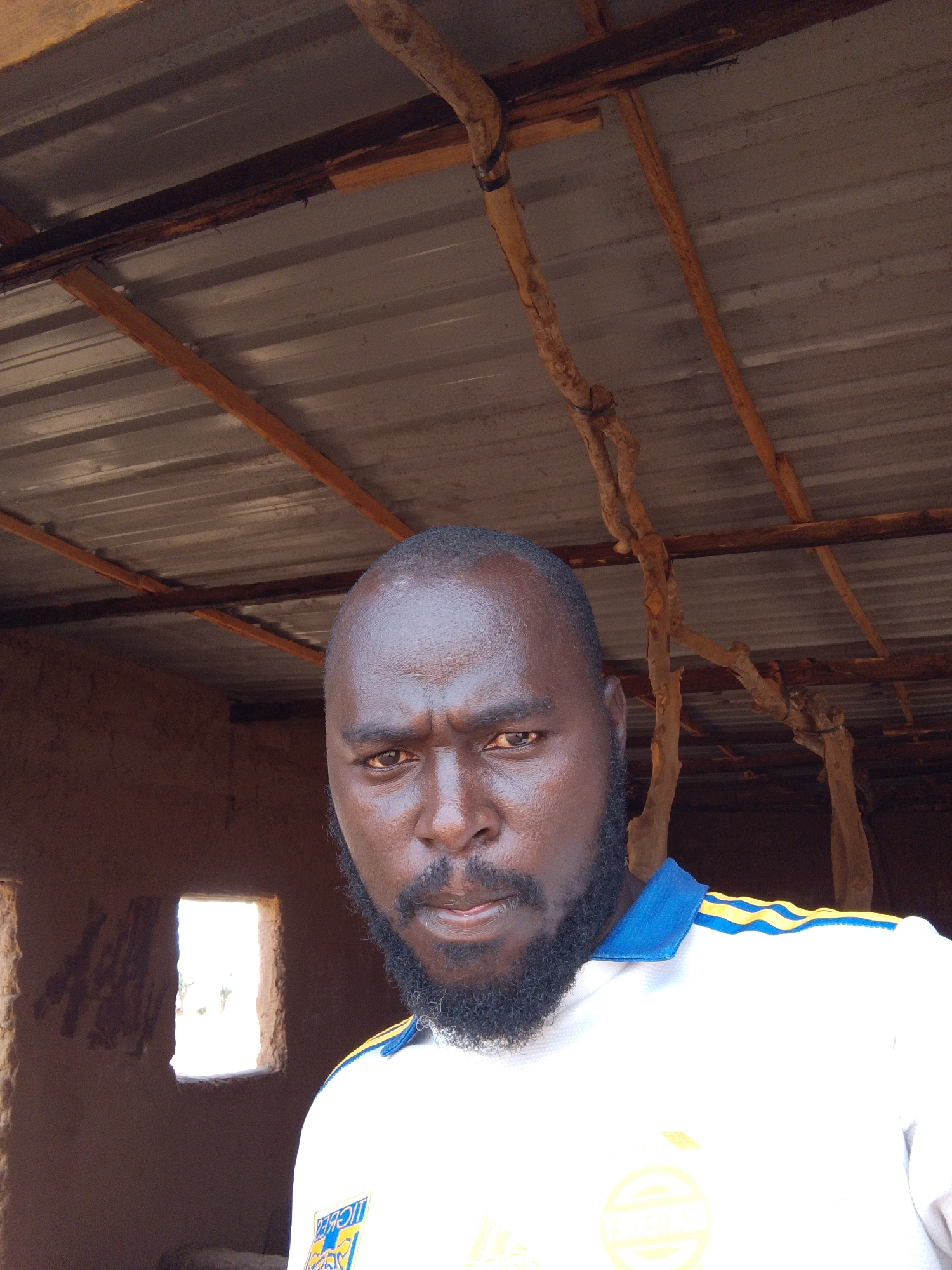 A man with dark skin, a shaved head, and a full black beard is centered in the frame, appearing to be indoors. He wears a white and blue collared shirt. Behind him, the structure appears to be made of mud and wood. A window is visible in the mud wall, letting in daylight. The ceiling is corrugated metal supported by wooden beams. Based on the prompt the photo was taken in Mulke Barikin Sayejo, Nigeria.