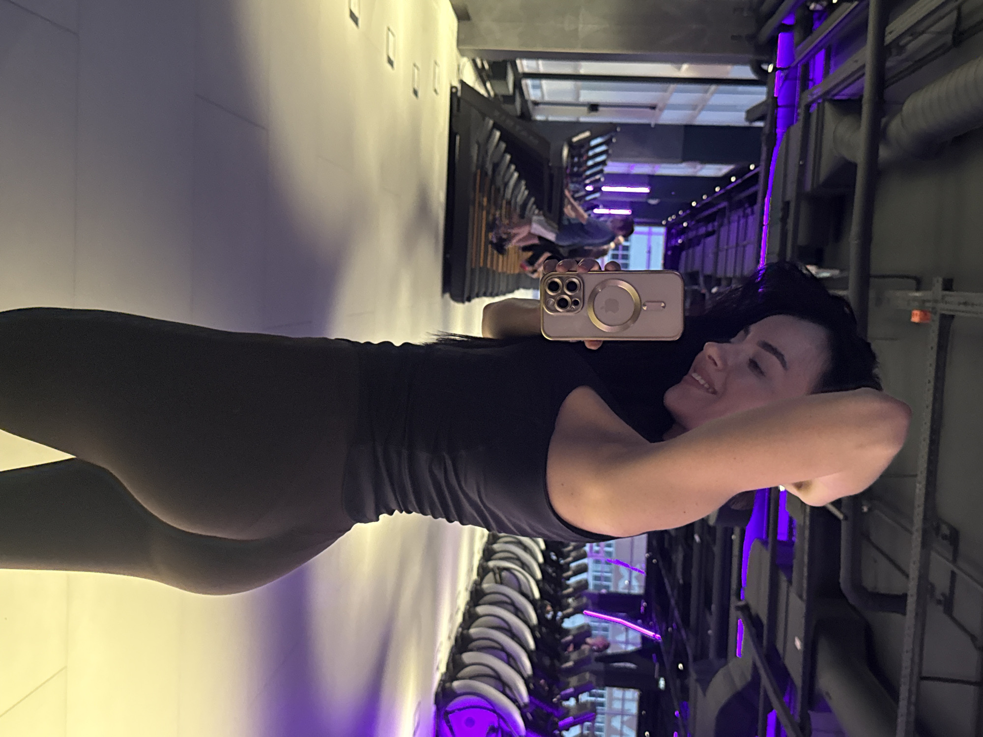 A woman wearing dark leggings and a tank top is taking a selfie in what appears to be a fitness studio or gym. The studio has modern, industrial-style architecture with exposed metal beams and pipes. The gym is illuminated by purple neon lights. A mirrored wall shows people working out on treadmills.