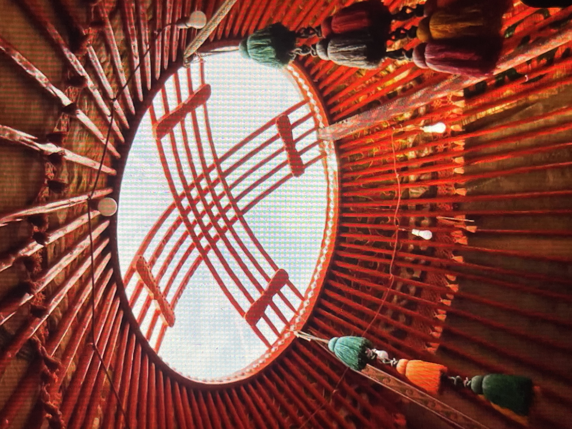 This media file captures the interior of a traditional Kyrgyz yurt, looking up towards the central opening, known as the *tunduk*. The scene is bathed in the bright, diffused light filtering through the *tunduk*, suggesting it is daytime, possibly with clear skies. The structure of the yurt is prominently displayed, with wooden lattice walls (*kerege*) made of thin, vertically oriented poles painted red, forming a circular base. Extending inwards and upwards from the *kerege* are angled wooden poles forming the roof structure (*oonok*), which converge towards the *tunduk*.
The *tunduk* itself is a circular opening covered with a transparent material, allowing the sky to be visible. It is framed by a red wooden lattice crisscrossed with additional red wooden beams. Dangling from the ceiling and walls are decorative tassels in various colors, including green, orange, and maroon, adding a vibrant touch to the rustic interior. A few bare lightbulbs are also visible, indicating the presence of artificial lighting.
The overall impression is one of a cozy and traditional living space. The dominant color palette is red, from the wooden structure, contrasted with the earthy tones of the yurt's felt or fabric lining and the bright colors of the decorative elements. There is no visible text in the media file. The image conveys a sense of cultural authenticity and the unique architectural style of Central Asian nomadic dwellings. The focus is on the structural elements and decorative details, without any people present.