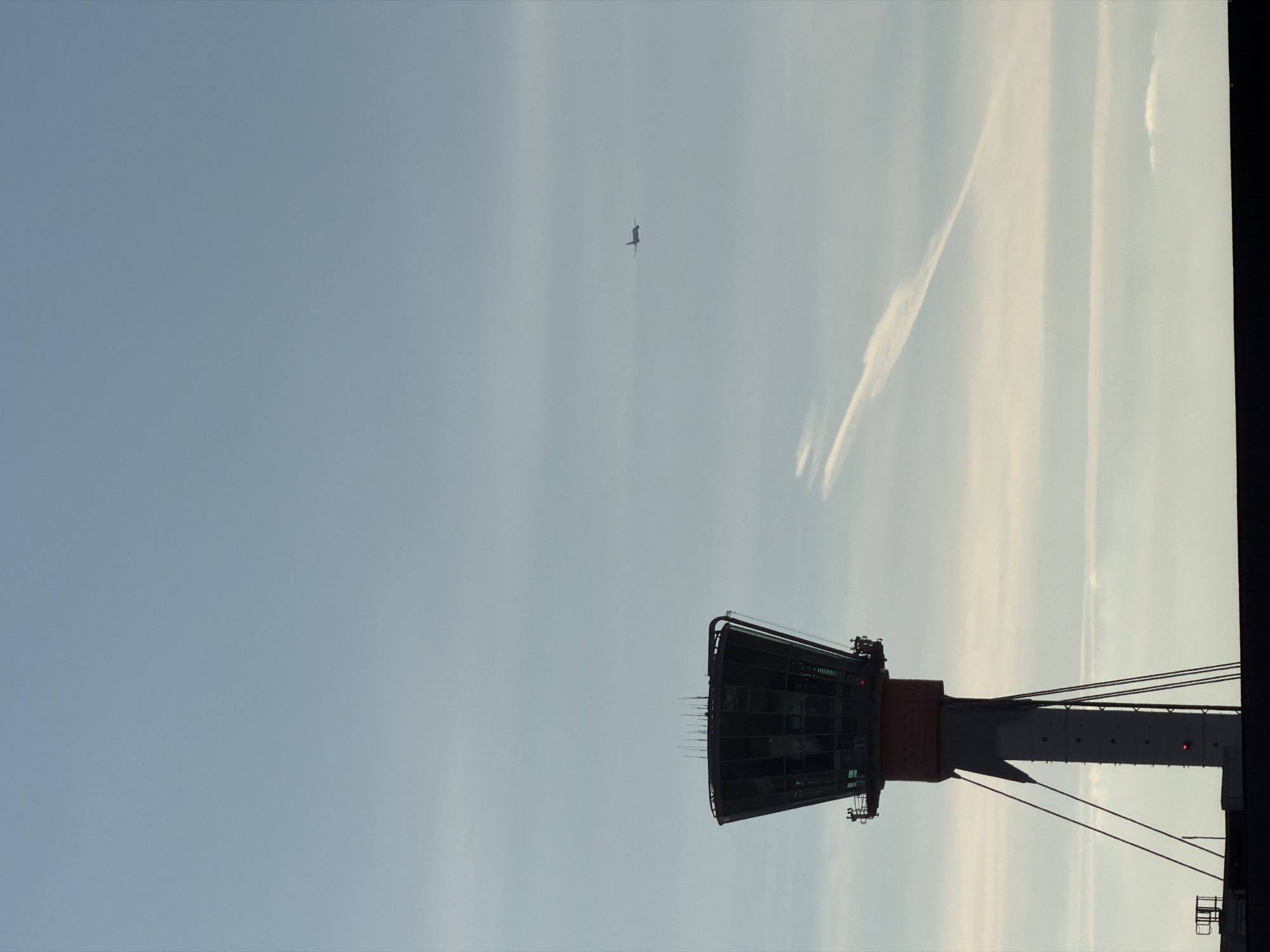 The image shows a daytime view of the control tower at London City Airport (LCY) in London, United Kingdom. An airplane is visible in the distance, in the upper center of the frame. Contrails are also visible in the sky. The image is taken from a low angle, looking upwards toward the control tower and the sky. The right side of the image is somewhat obscured by a darker object.