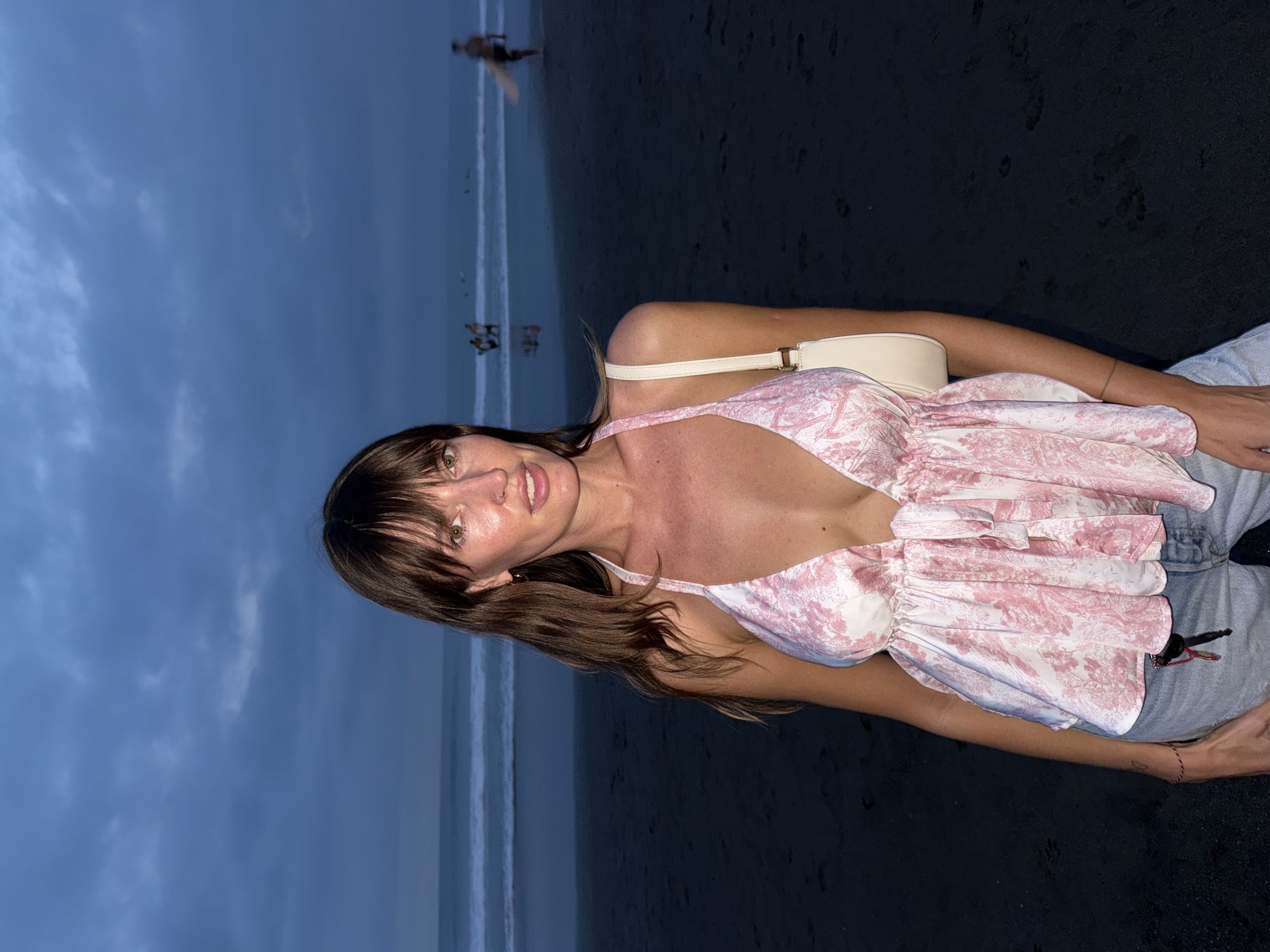 A medium-shot, rotated, low-angle photo shows a fair-skinned woman with brown hair and bangs, wearing a pink and white printed top, a white shoulder bag, and blue jeans, standing on a dark sand beach. Her hair is loose, and she is looking at the camera with a slight smile. The background includes the ocean at dusk with gentle waves, a cloudy sky, and a few distant figures on the beach. The dark sand shows footprints, suggesting recent activity. The photo appears to have been taken at a beach in Canggu, Indonesia, given the dark sand.