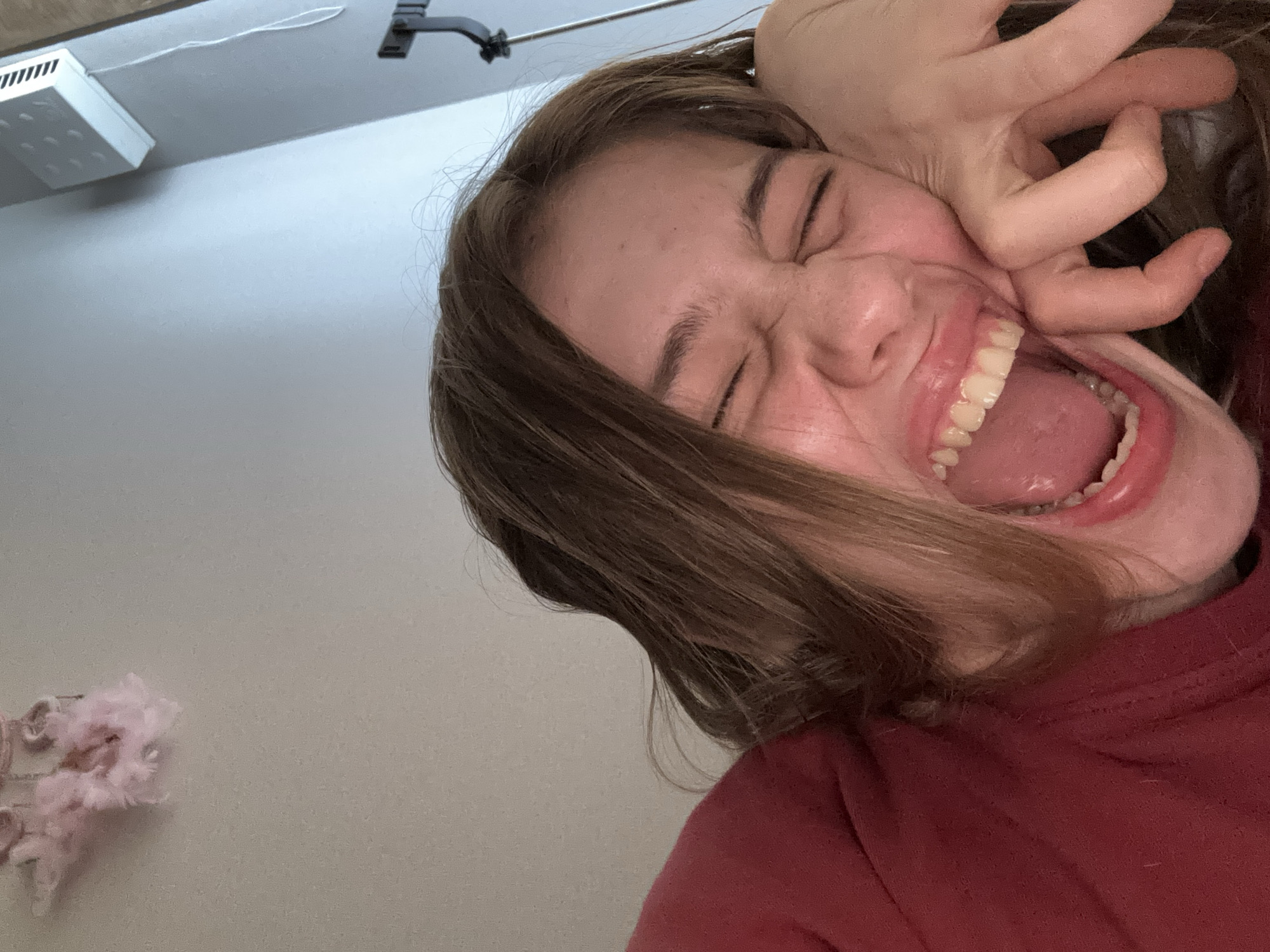 This is a close-up, low-angle selfie of a young person with brown hair, wearing a red shirt, who appears to be laughing or yawning widely with their eyes closed and hand near their face. The background consists of a light-colored wall and a glimpse of a grey wall with a white electronic device mounted on it, along with a black metal fixture. The image does not contain any information that would allow for its location to be determined, including in the city of Vsevolozhsk, Russia.