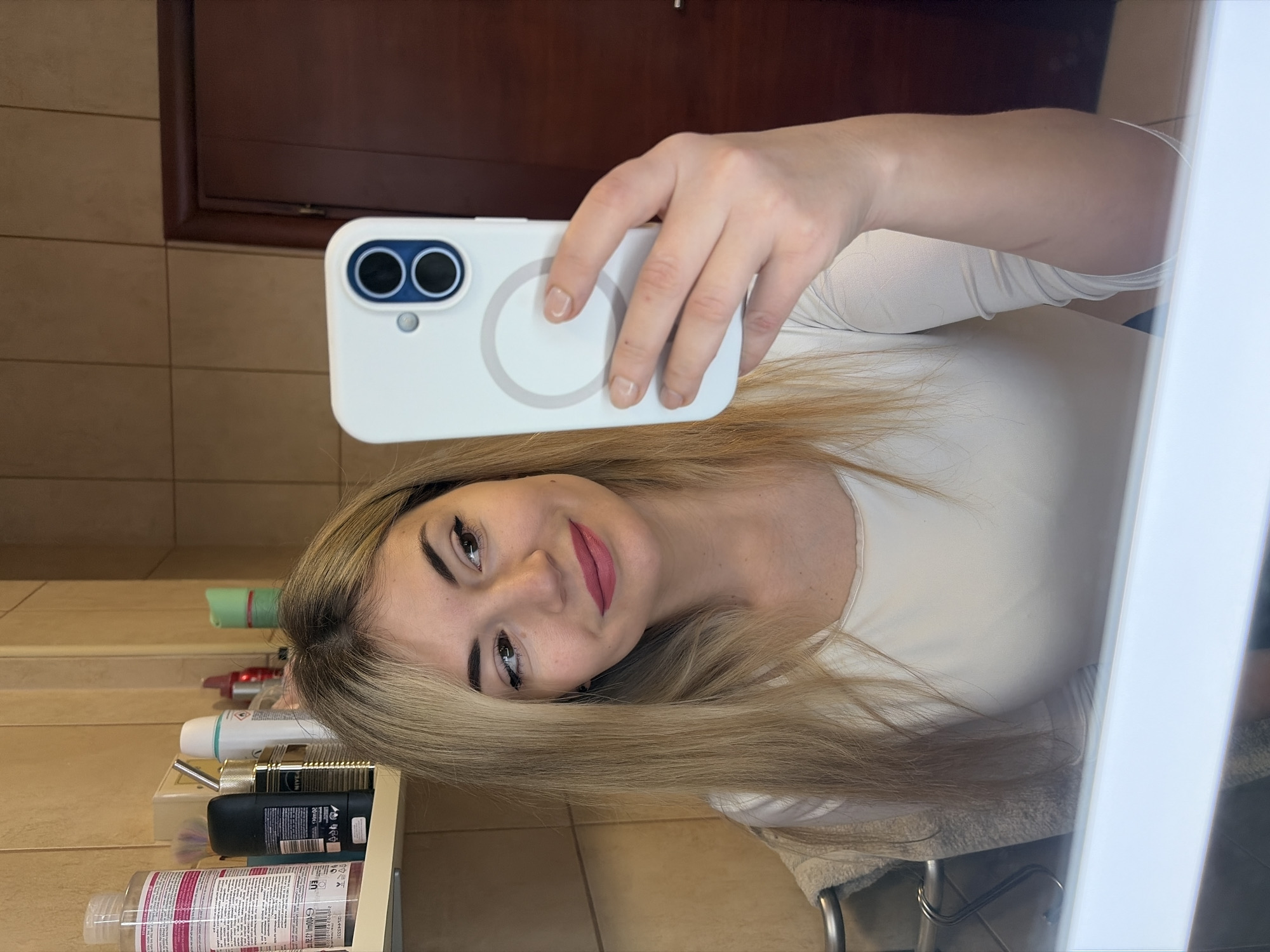A woman is taking a selfie in a bathroom. She is holding a white smartphone in her right hand. The background shows tiled walls and a dark wood door frame, along with toiletries on a shelf. A partial view of a mirror frame is visible on the right. There are no indications of this location being in Budva, Montenegro.