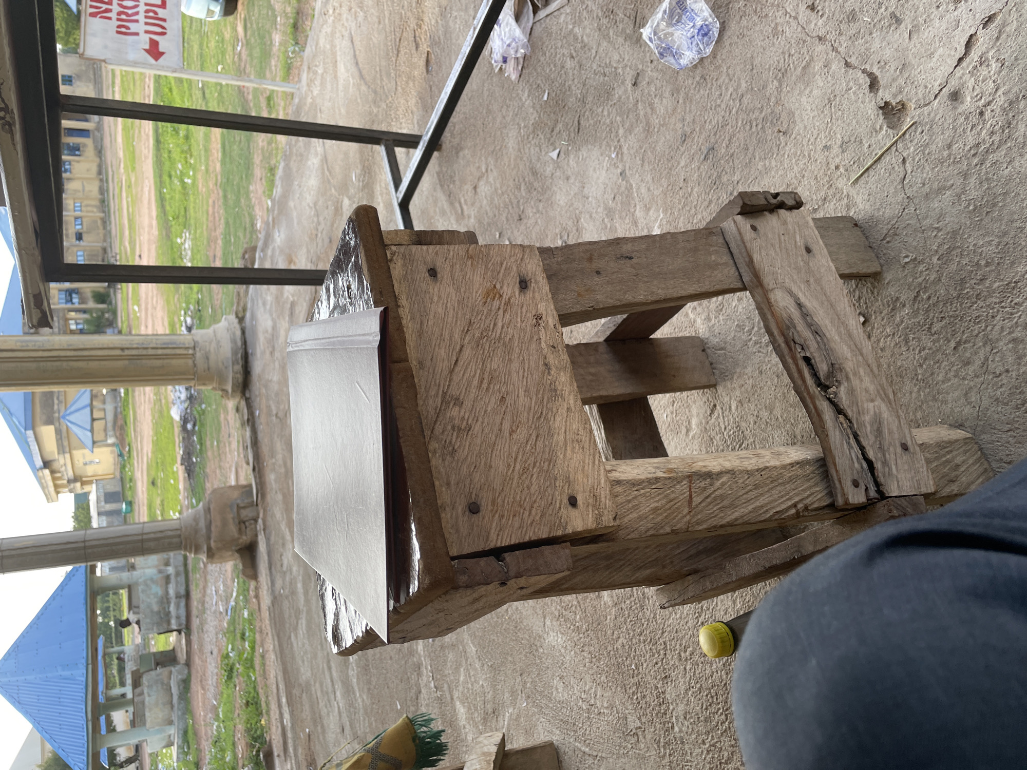 The image shows an outdoor scene in Jalingo, Nigeria. In the foreground, a weathered, rustic wooden stool sits on a concrete ground. A dark-colored book is placed on top of the stool. To the left of the stool, a person's dark blue pants are visible, and a yellow bottle cap is on the ground. In the background, there are some buildings with blue roofs, a metal structure, and dry, patchy grass. A red and white sign with text, partially obscured, is visible in the background. Scattered debris is present on the ground.
