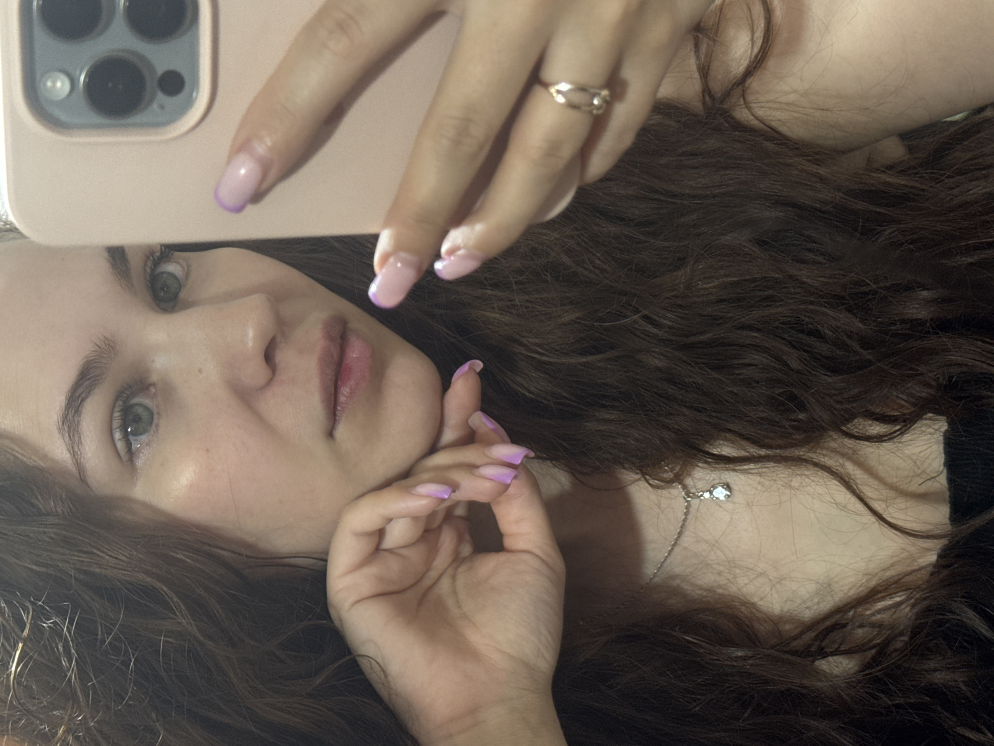 A young woman is captured in a close-up selfie, taken from an overhead perspective while she lies down. Her face is turned towards the camera, revealing light-colored eyes, defined eyebrows, and lips with a natural pink tone. Her long, dark brown, wavy hair is spread out around her head and shoulders, covering much of the background.
She holds a light pink or beige smartphone with a visible multi-lens camera module in her right hand, positioned above her head. Her nails are long and manicured with a light pink to purple ombré design. On her right ring finger, she wears a simple gold-toned ring. Her left hand is also visible, resting near her chin, showcasing similar ombré nails. A delicate silver-toned necklace with a single clear pendant is visible against her skin, nestled amongst her hair and a dark top.
The lighting is soft and even, suggesting an indoor setting without direct sunlight. Her expression is calm and neutral, looking directly into the phone's camera. No text is visible in the image. The scene takes place indoors, potentially in a residence in Chaikovsky, Russia.