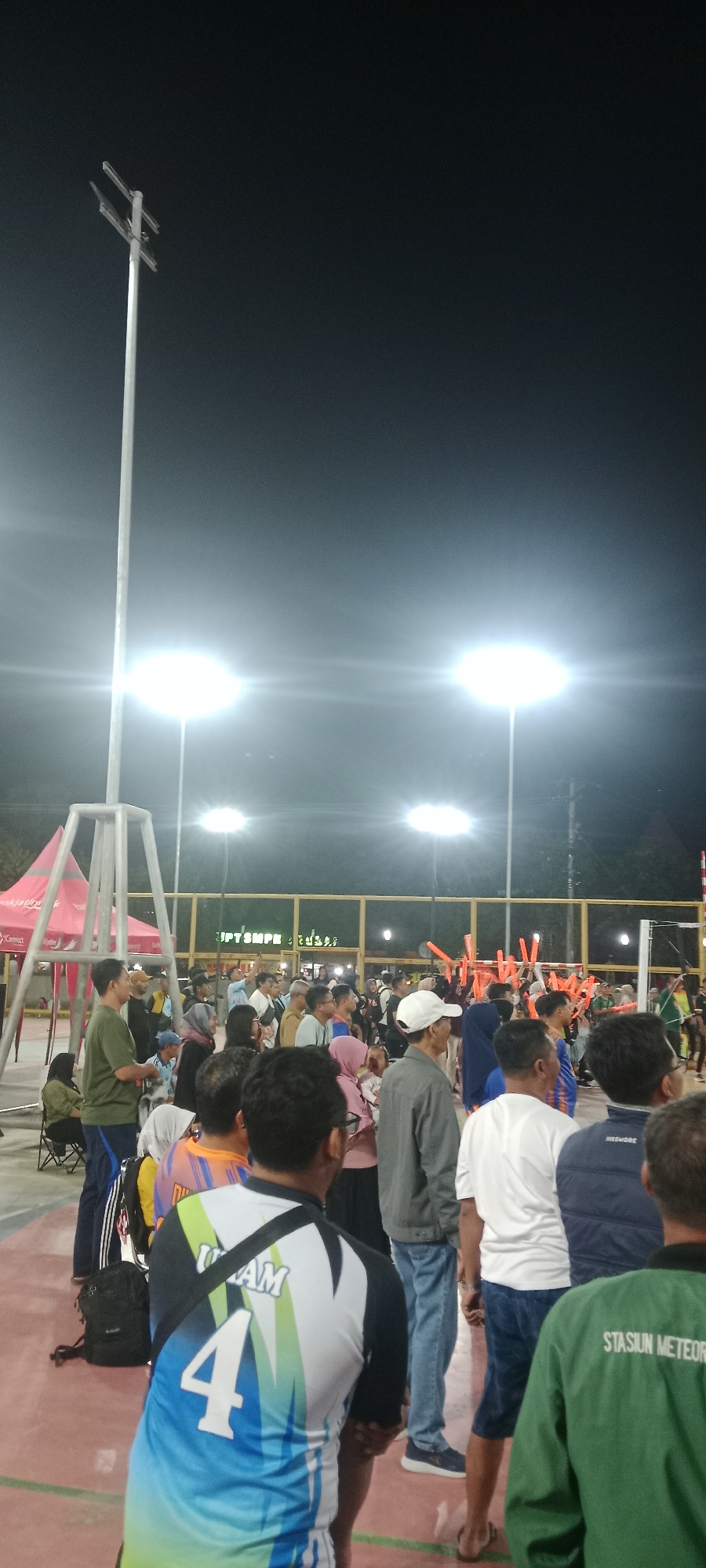 The image captures an outdoor gathering at night, likely a sporting event or public assembly, at the Gelora Hayam Wuruk field in Tuban, Indonesia. Bright floodlights illuminate the scene, creating a stark contrast with the dark sky. A crowd of people, predominantly adults, are visible, many standing and observing an event or activity not fully depicted. Some individuals are wearing athletic attire, including jerseys with numbers and logos, suggesting a sports context. There's a volleyball net in the background, further supporting this interpretation. Several orange noisemakers are held by people in the crowd, indicating an atmosphere of excitement or support. A red tent with the "Connert" logo is partially visible on the left, possibly serving as a vendor or information booth. The ground appears to be a court, consistent with a sports venue. The overall environment is public and well-lit for nighttime activity.