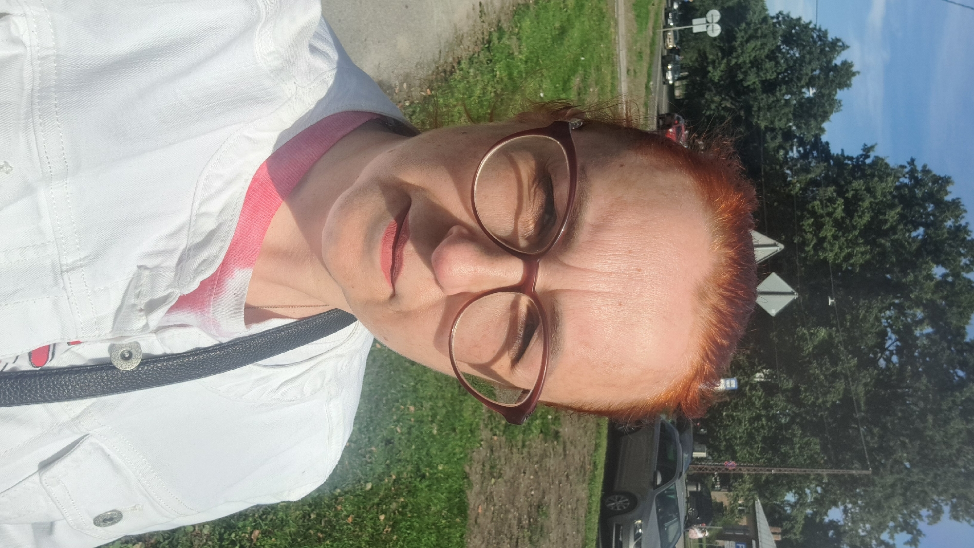A selfie of a middle-aged woman with red hair and glasses, outdoors in a city setting. She is wearing a white denim jacket over a pink and white tie-dye t-shirt and a simple silver necklace. The background shows a tree-lined street with cars, traffic signs, and buildings. The image appears to be taken on a sunny day. The woman is looking directly at the camera with a neutral expression. The context of Riga, Latvia is not discernible from the visual information in the image.