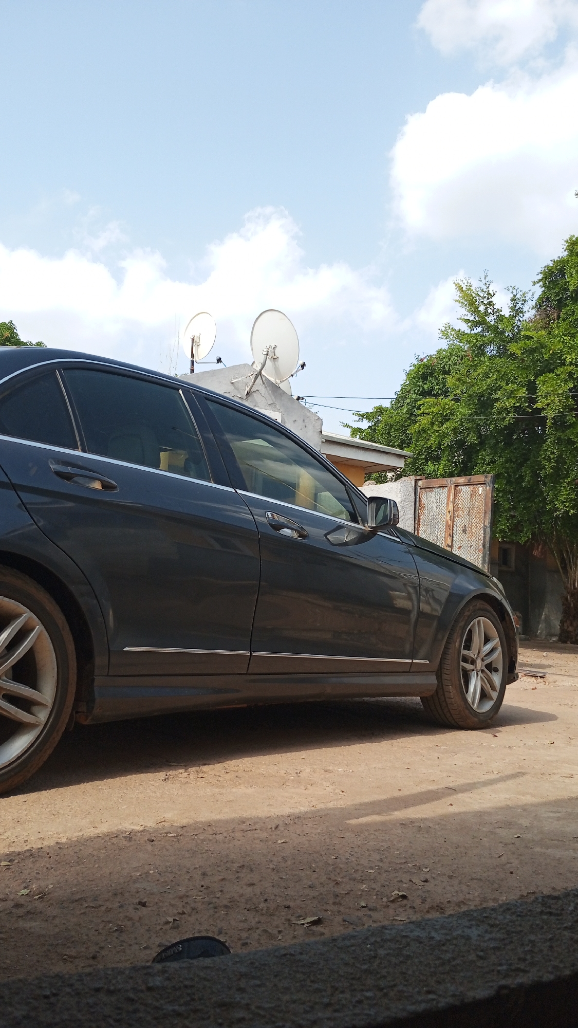 The image shows a dark-colored Mercedes-Benz C-Class sedan parked on a dirt road or yard in Adankolo, Nigeria. The vehicle is facing right. In the background are a building with two satellite dishes on the roof, trees, and a metal gate or door. The sky is blue with scattered clouds. The scene appears to be during daylight. No people are visible.