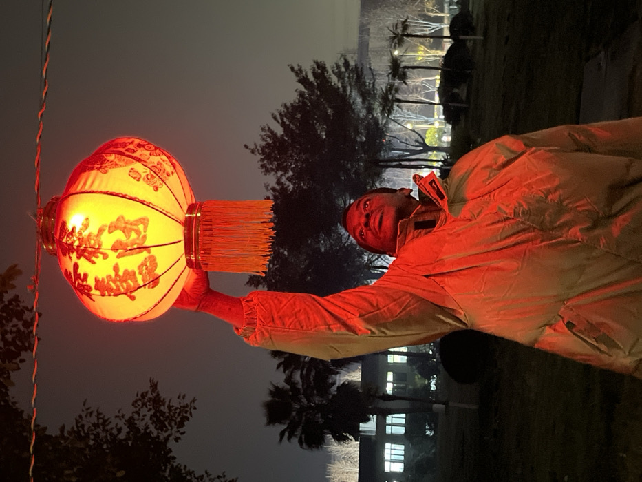The image depicts a nighttime scene in Jiangjin, China. A dark-skinned individual, appearing male, is positioned centrally, illuminated by a vibrant red glow. They wear a light brown puffer jacket. Their right arm is raised, holding a large, illuminated spherical red Chinese lantern directly above their head. The lantern features yellow-gold Chinese characters and red tassels hanging from its base. The individual's face, bathed in red light, is directed upward and slightly to the left. The background is dark, revealing silhouettes of trees, including palm trees, and a multi-story building with illuminated windows in the mid-ground. Distant artificial lights are visible on the right side, suggesting an urban or semi-urban park-like environment with grass and various foliage. A wire, potentially part of a lantern string, is visible in the upper left.