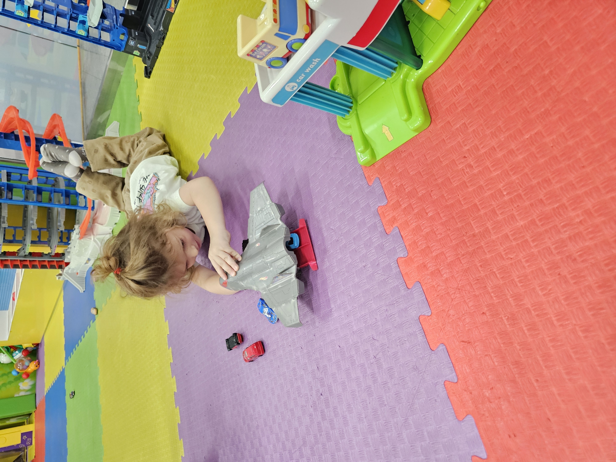 A young child lies on a colorful foam play mat in what appears to be an indoor play area. The mat is composed of interlocking squares in shades of red, purple, yellow, green and blue. The child has curly blonde hair pulled back in a ponytail with a red scrunchie, and is wearing a white t-shirt and brown pants. They are engaged in playing with toy cars and a gray toy airplane. A toy car wash is positioned nearby, at the edge of the playmat. The location is in the city of Khimki, Russia. Other toys, including a blue shelving unit holding various items, are visible in the background.