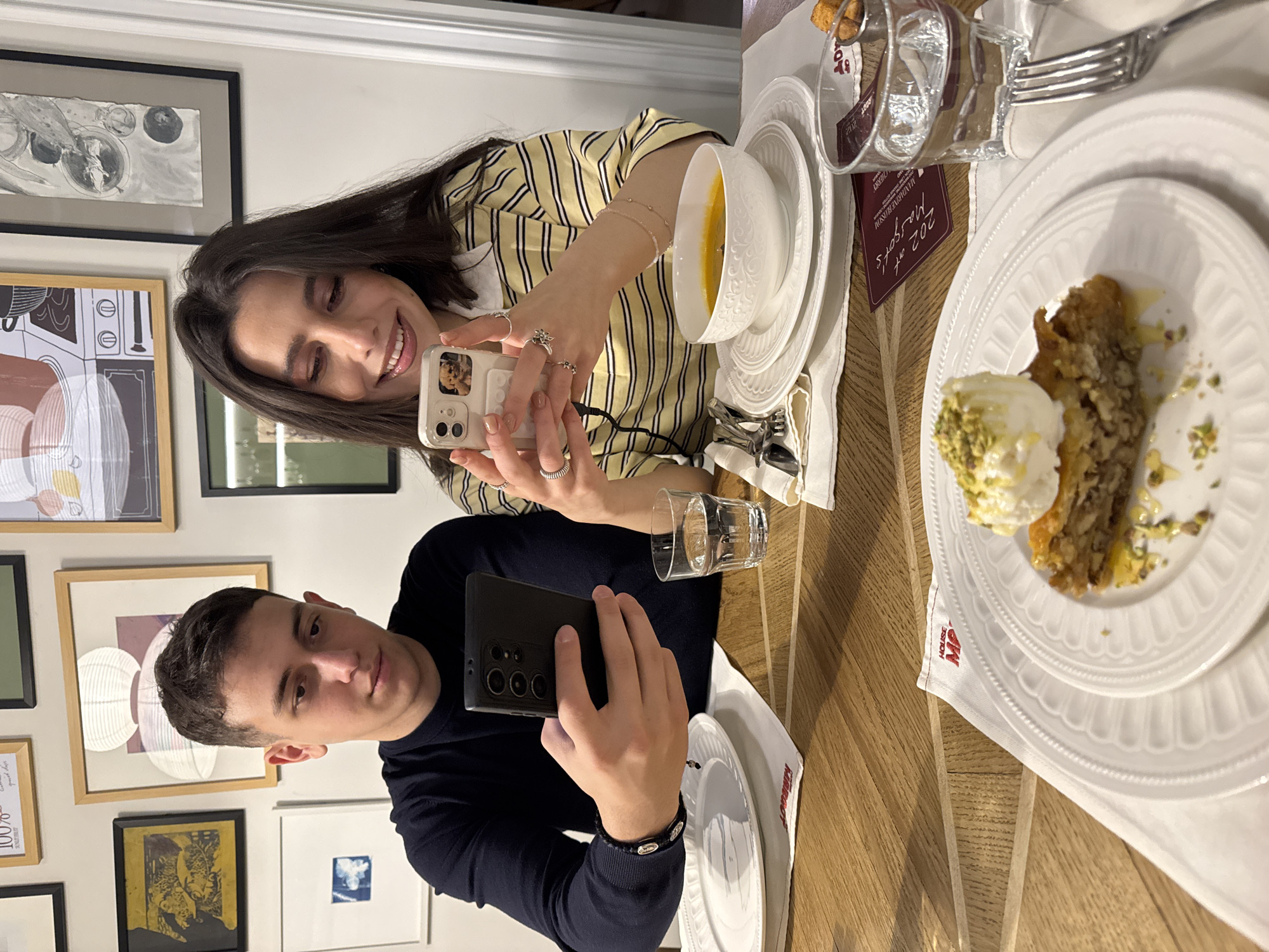 The image shows two people sitting at a table, likely in a restaurant, and taking pictures of food. A woman in a yellow and white striped shirt is taking a picture with her phone, while a man in a navy sweater is also taking a picture with his phone. There is a bowl of soup on a plate to the right of the woman and a plate of baklava with ice cream to the right of the man. There is a glass of water next to each plate. Based on the "House MP" logo on the napkin, the "MANDARINE RUSCONI" card, and the style of the restaurant, the location is likely Mandarine Rusconi restaurant in Yerevan, Armenia. The walls have multiple framed pictures.