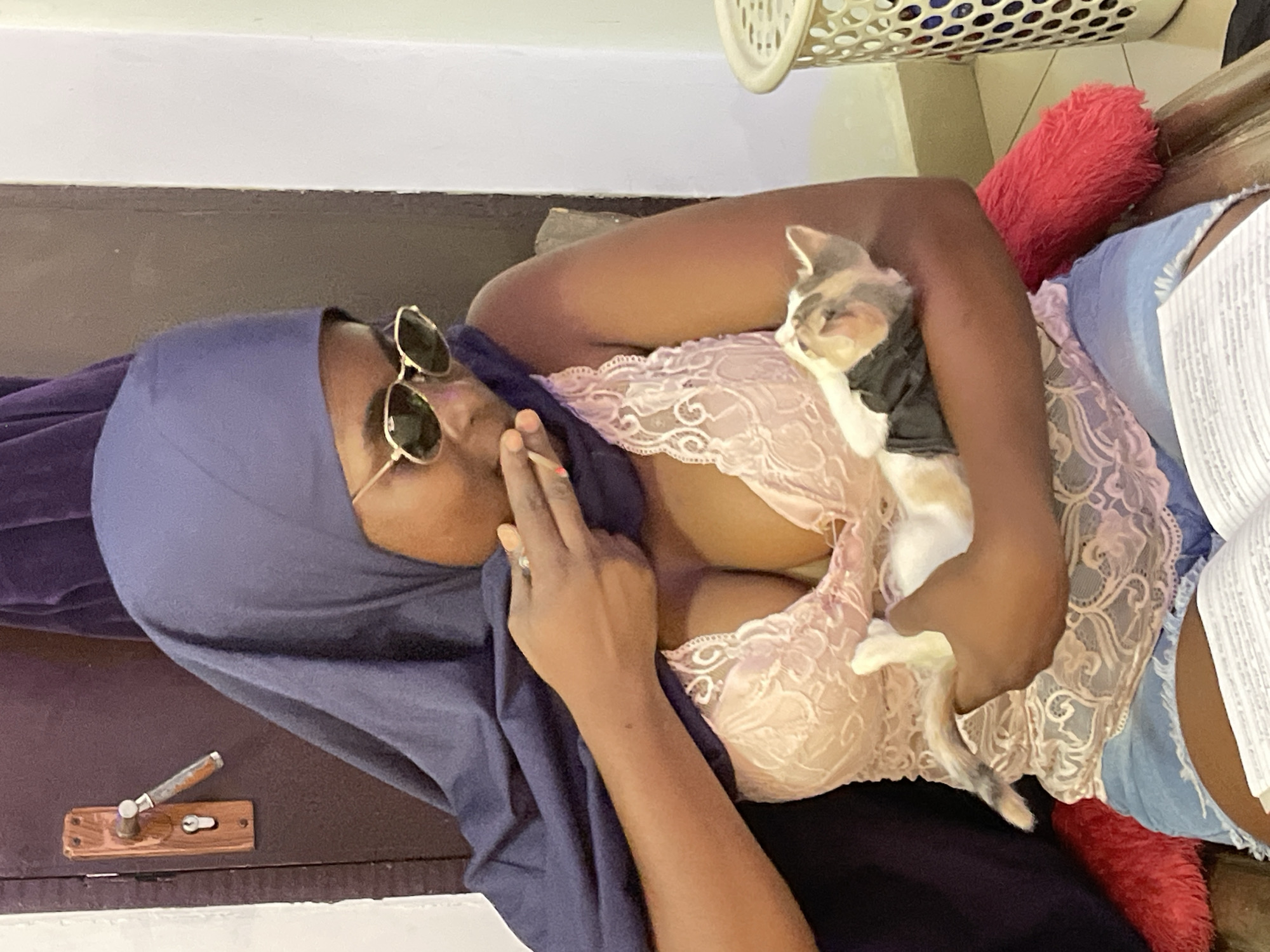 A woman is lying down, wearing a blue hijab and sunglasses, with a light pink lace bra visible. She is holding a cat wearing a dark-colored shirt. A red pillow and a woven plastic basket are visible in the background. There is a wooden door with a metal handle and lock. It is impossible to confirm the location is in Gwarinpa, Nigeria.