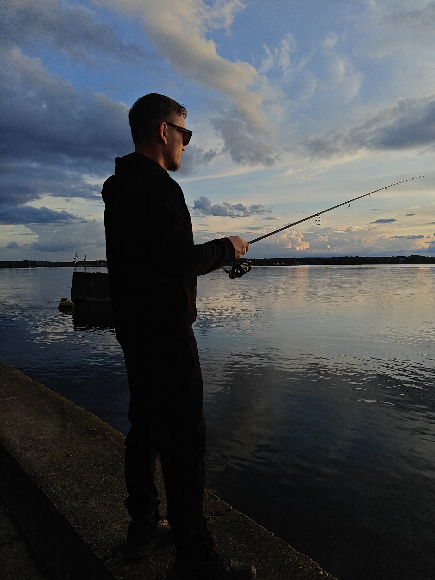 A man is fishing at sunset. He stands on a concrete pier, wearing sunglasses and a dark hoodie. He holds a fishing rod in his hands, casting the line out into the water. In the background is a body of water, with a shoreline visible in the distance. The sky is filled with clouds, colored by the setting sun. The location is Chaikovsky, Russia.