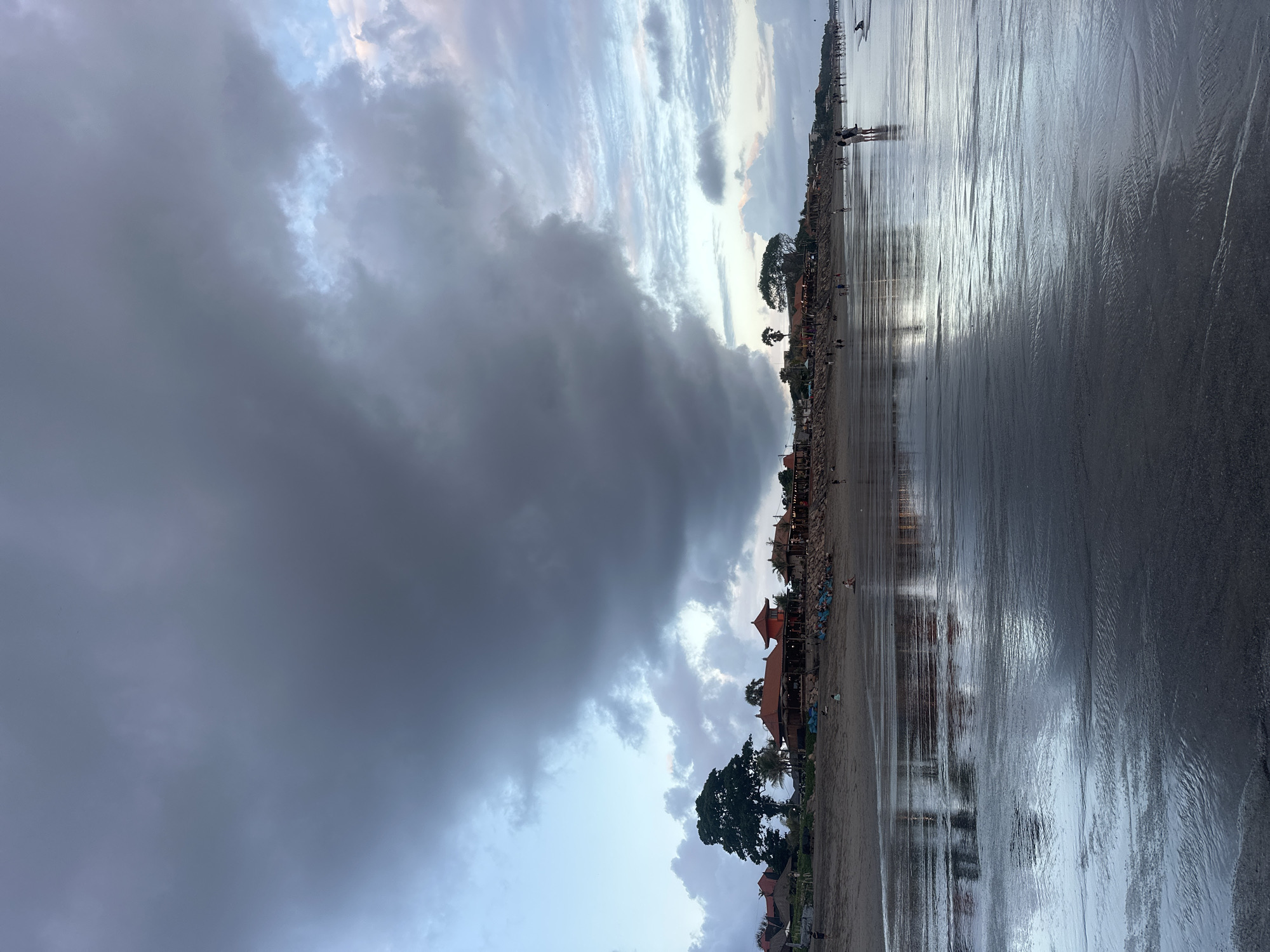 An eye-level, full shot captures a wide, wet sand beach in North Kuta, Indonesia, at dusk. The ocean's surface reflects the moody, overcast sky, with heavy, dark clouds dominating the upper half of the frame. The horizon line is low, with a strip of sand bordering a line of buildings, some with red-tiled roofs, set back from the beach. A large, dark green tree stands prominently on the left side of the frame. A few scattered individuals and groups of people are visible on the beach and near the buildings, some walking and others sitting. The wet sand in the foreground is smooth and reflective, with subtle ripples indicating receding water. The overall atmosphere is somber, yet peaceful, with the dim light suggesting the end of the day.
