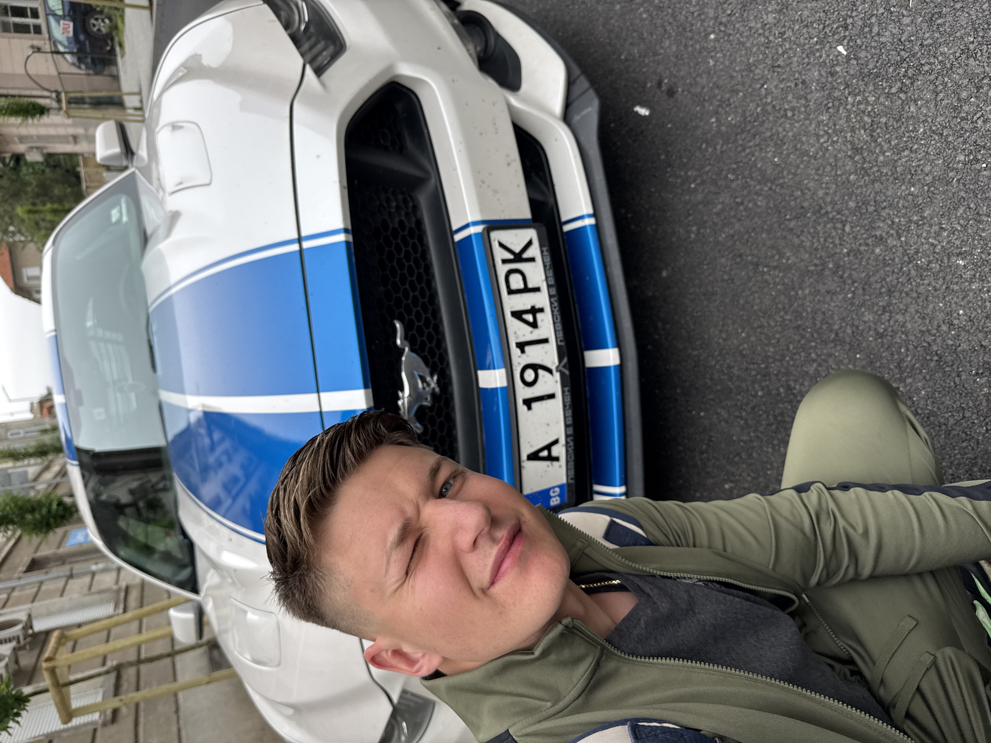 A close-up shot depicts a man with a short haircut wearing a green tracksuit posing next to a white and blue Ford Mustang car with a Bulgarian license plate A 1914 PK. The man is winking at the camera. The location is potentially in Burgas, Bulgaria, based on the license plate format. The background shows a street scene with buildings and other vehicles. The image is taken at ground level, presenting a slightly upward angle towards the car and the man.