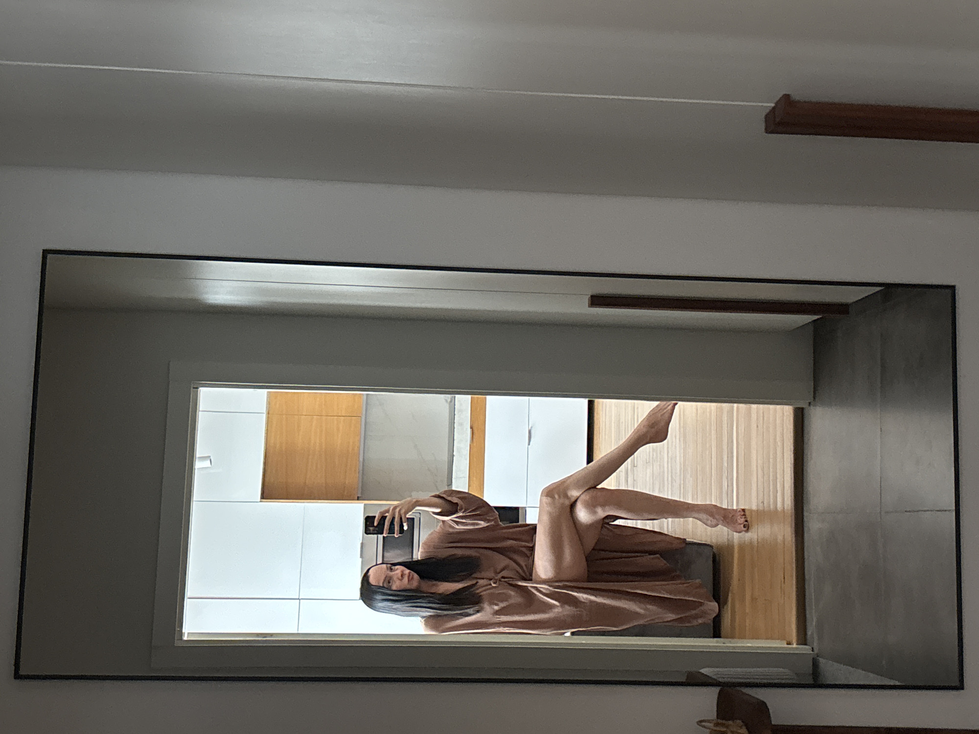 A mirror reflects a woman in a brown robe reclining on a gray chair, holding a phone. Behind her, the reflection shows a kitchen area with white cabinets and wooden accents, and a hallway with a wooden floor. The environment outside the reflection includes parts of the room with a ceiling, wall, and trim. There is no evidence to support the location is in Chaikovsky, Russia.