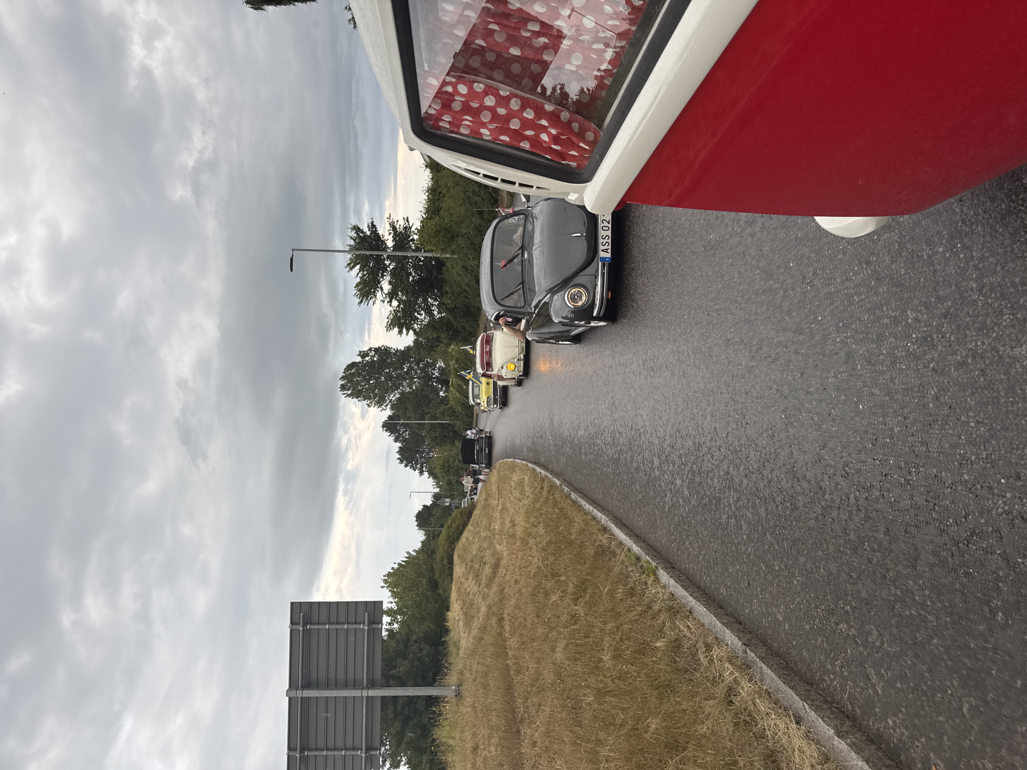 The image shows a procession of vintage Volkswagen cars driving on a wet asphalt road. In the foreground, a red and white vehicle, likely another vintage Volkswagen, is partially visible from the side. Several Volkswagen Beetles are visible in the procession, with one dark grey Beetle in the middle ground displaying the license plate "ASS 02". Yellow and white Beetles are also part of the line. The road is bordered on the left by a grassy embankment and on the right by trees. The sky is overcast and grey. The scene appears to be on a highway or a main road, possibly near the city of Falkenberg, Sweden, as suggested by the presence of Swedish flags on some of the vehicles and the overall landscape. The cars are driving in the same direction, indicating a parade or a group outing.