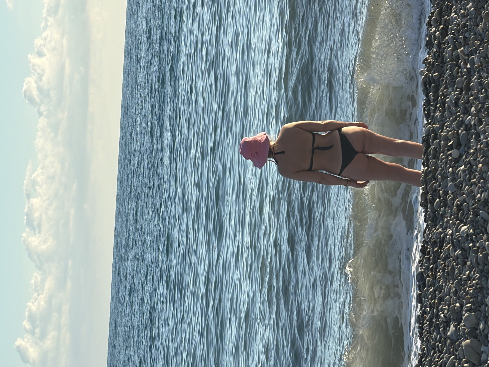 A woman wearing a pink bucket hat and a black bikini stands on a pebble beach, facing the sea. The waves are breaking gently on the shore. The sky is a pale blue with scattered white clouds. The image appears to have been taken during daylight hours, possibly late afternoon given the lighting. The location is identified as Sochi, Russia, implying a coastal setting typical of the Black Sea.