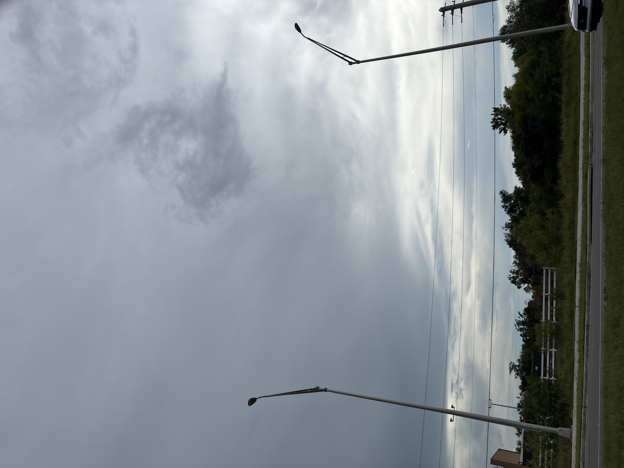 The image depicts an outdoor scene with a cloudy sky above a roadside environment. Two tall, slender streetlights are visible, one on the left and another on the right, both against the overcast sky. Below the sky, there are power lines stretching horizontally across the frame. The middle ground features a line of trees and vegetation, with a white fence visible behind some of the foliage. A patch of grass borders a paved road in the foreground. A portion of a white vehicle is visible on the far right of the image. The setting appears to be a suburban or semi-rural area. No people are present in the image. The lighting suggests it is daytime, possibly before or after rain due to the heavy cloud cover.
