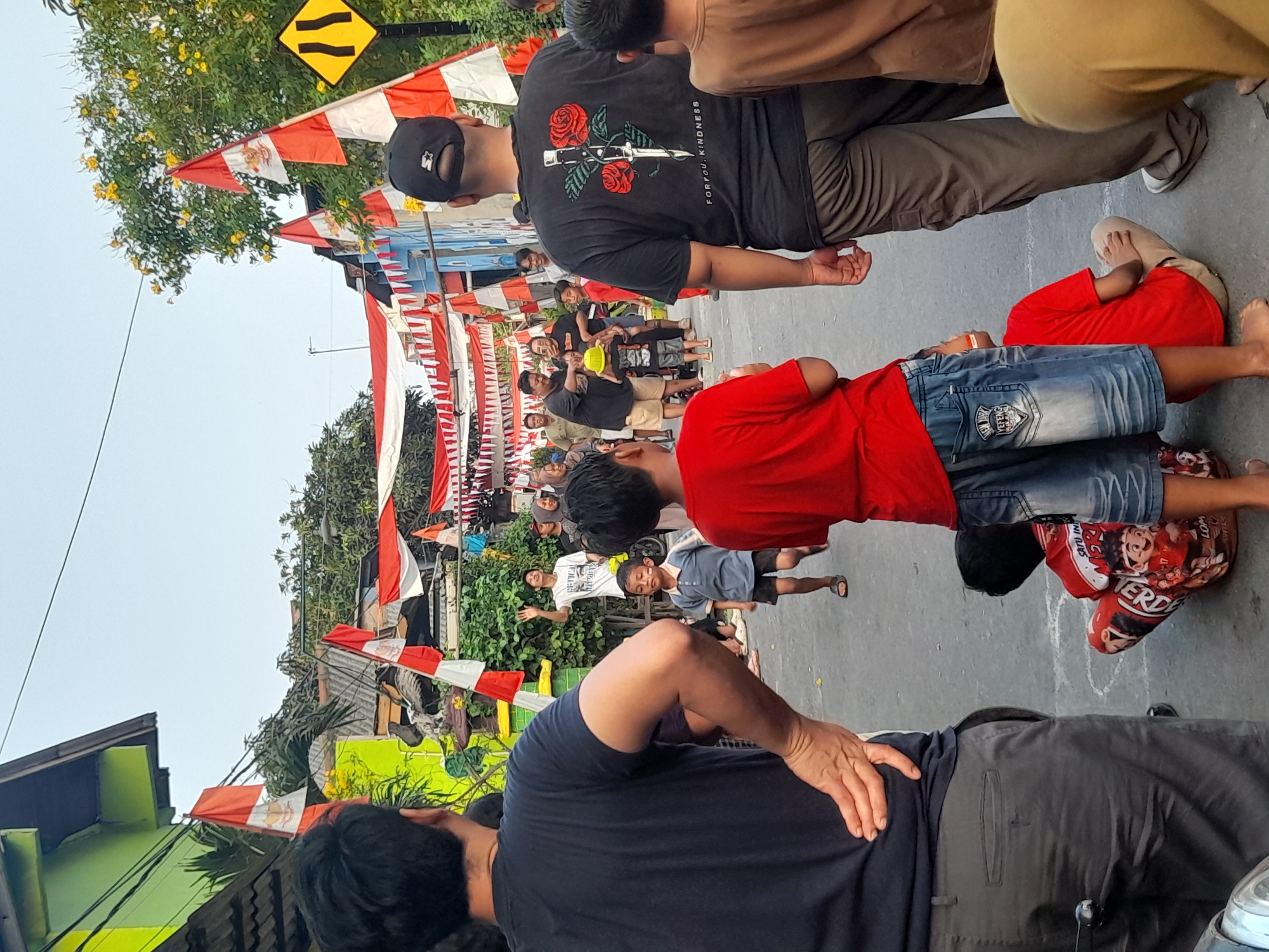 The image shows a street scene in North Jakarta, Indonesia, decorated with Indonesian flags and banners for a celebration. A group of people, including men, women, and children, are gathered in the street. Several children are playing or watching an activity, with one boy in the foreground wearing a red shirt and blue jeans, and another boy in a blue shirt and shorts running. Adults are standing and observing the children. Buildings line the street, with trees and greenery visible. The atmosphere appears festive.